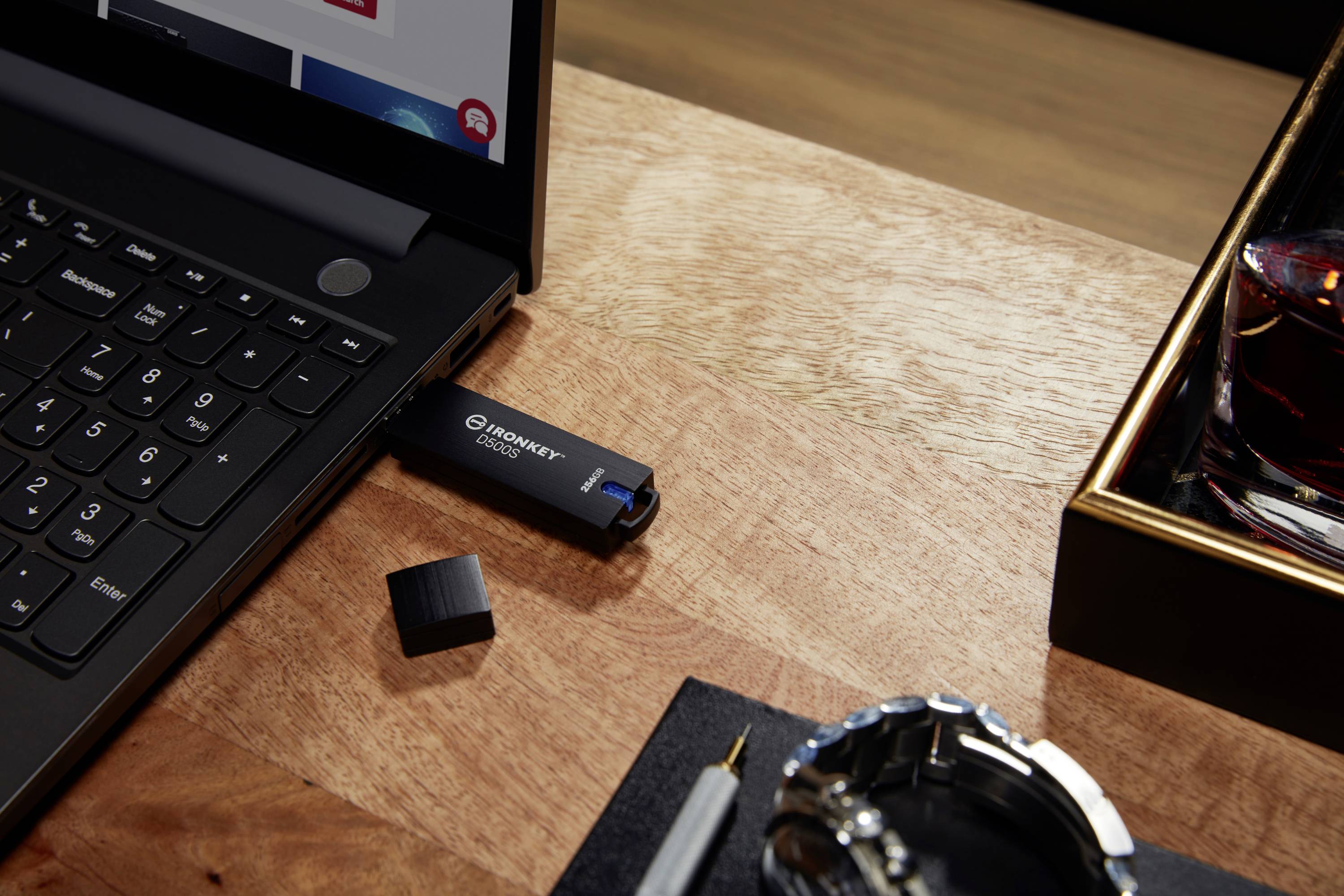 Een USB-stick steekt in een laptop op een houten tafel. Ernaast liggen een horloge, een pen en een glas.