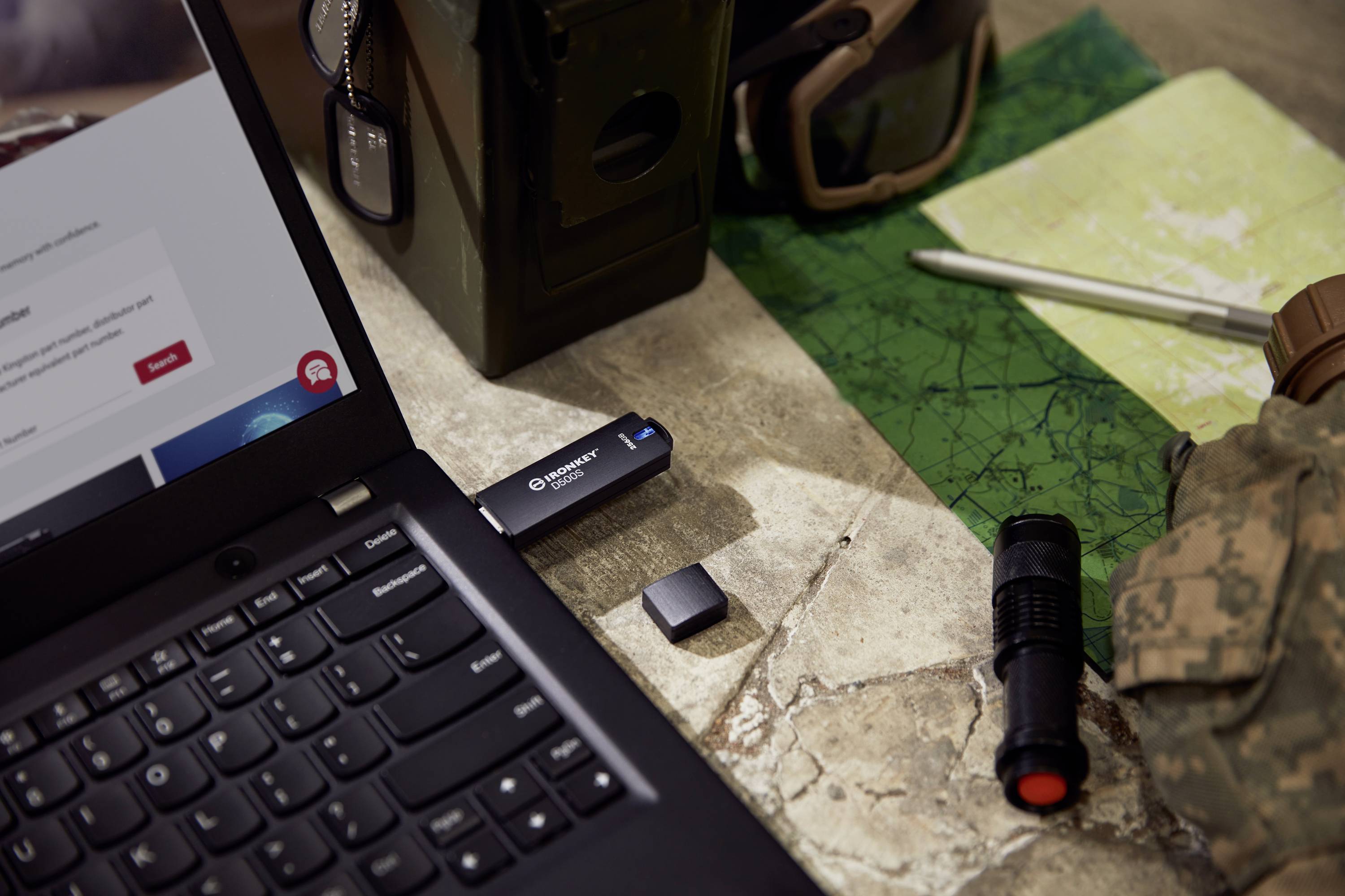 Laptop op tafel met USB-stick, kaart, zaklamp, bril en helm; symboliseert buitenactiviteiten of militaire planning.
