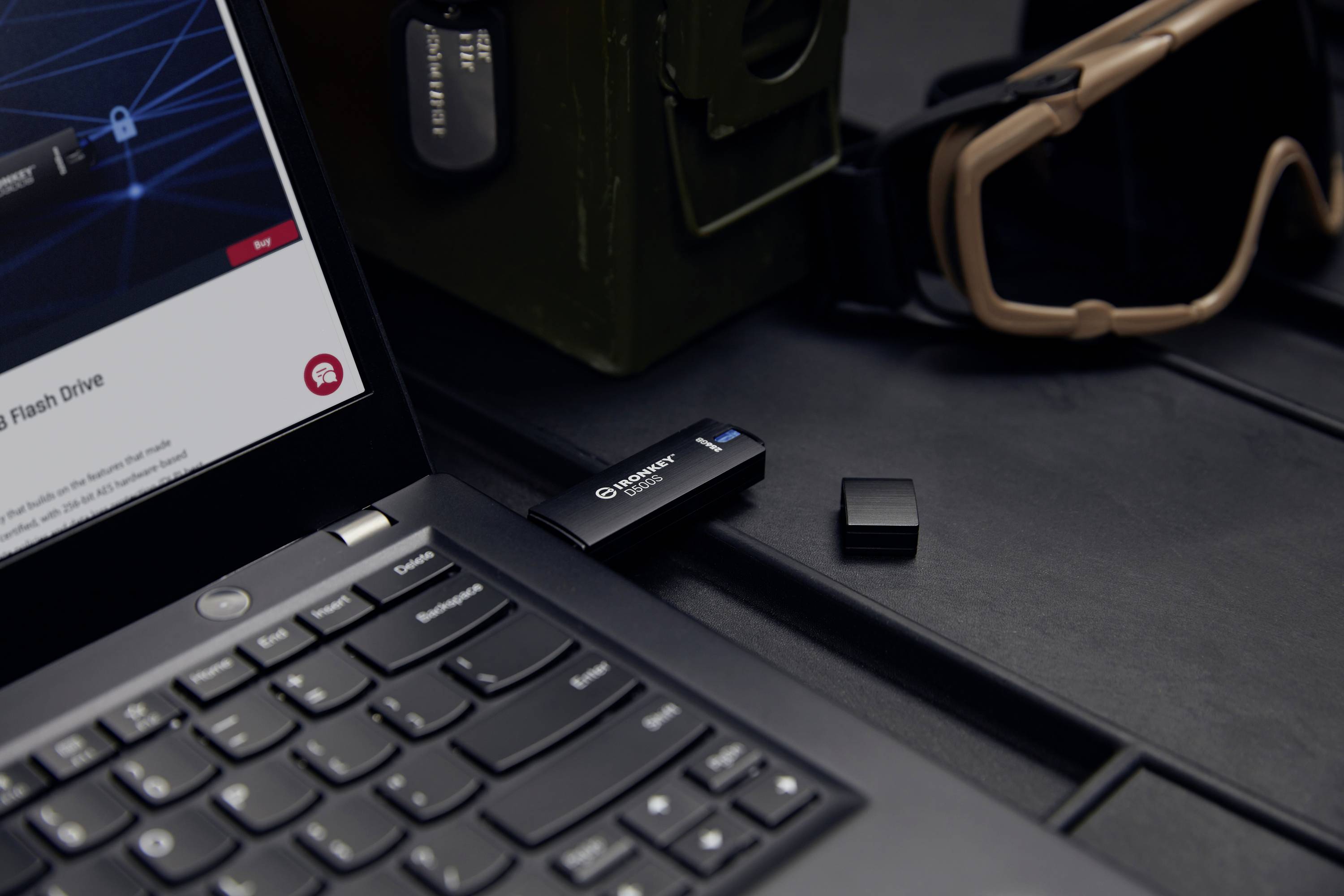 Een USB-stick steekt in een laptop. Naast de laptop liggen een dop en een veiligheidsbril. Een groene koffer op de achtergrond.