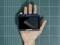 Een hand houdt een Arduino-touchscreen vast boven een groene, geruite achtergrond. Het scherm is uitgeschakeld.