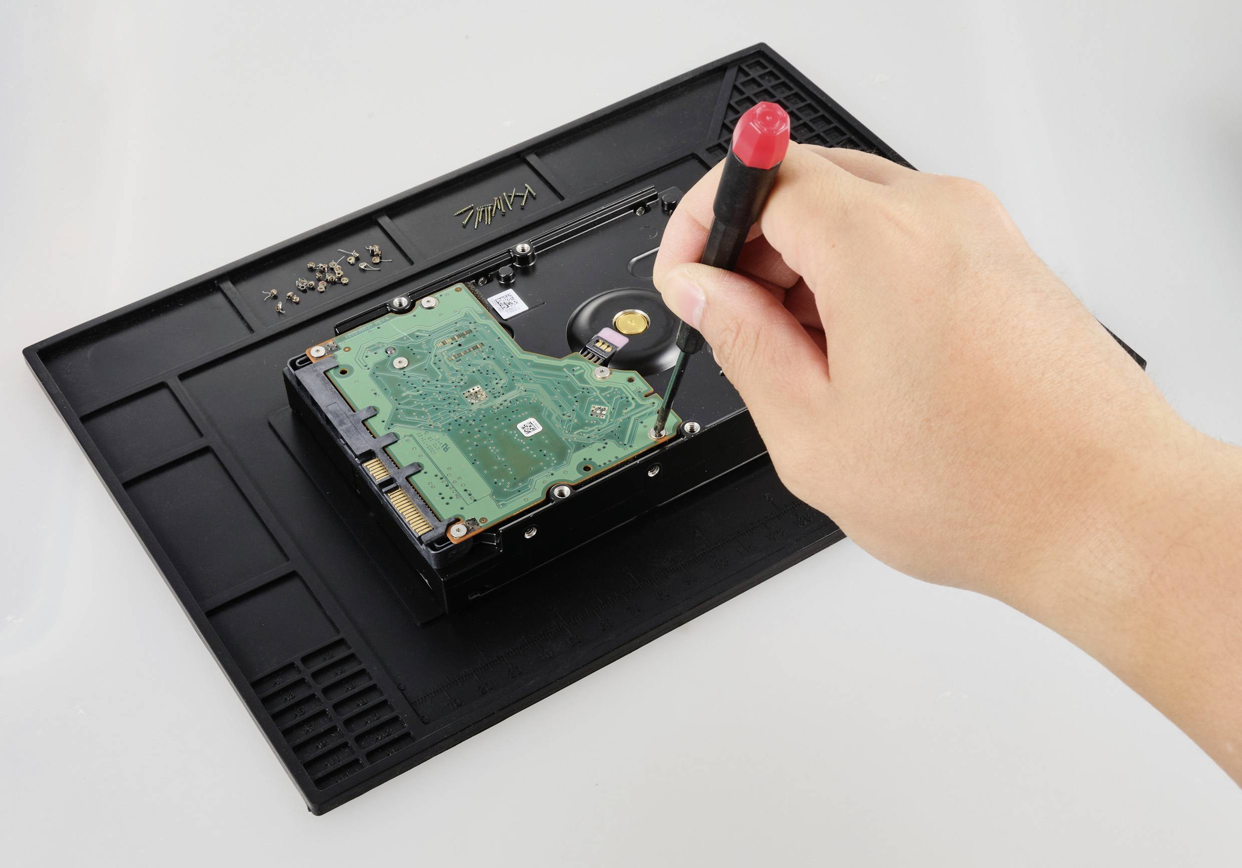 Een hand gebruikt een schroevendraaier om een harde schijf te repareren op een antistatische mat. Schroeven liggen in de buurt.