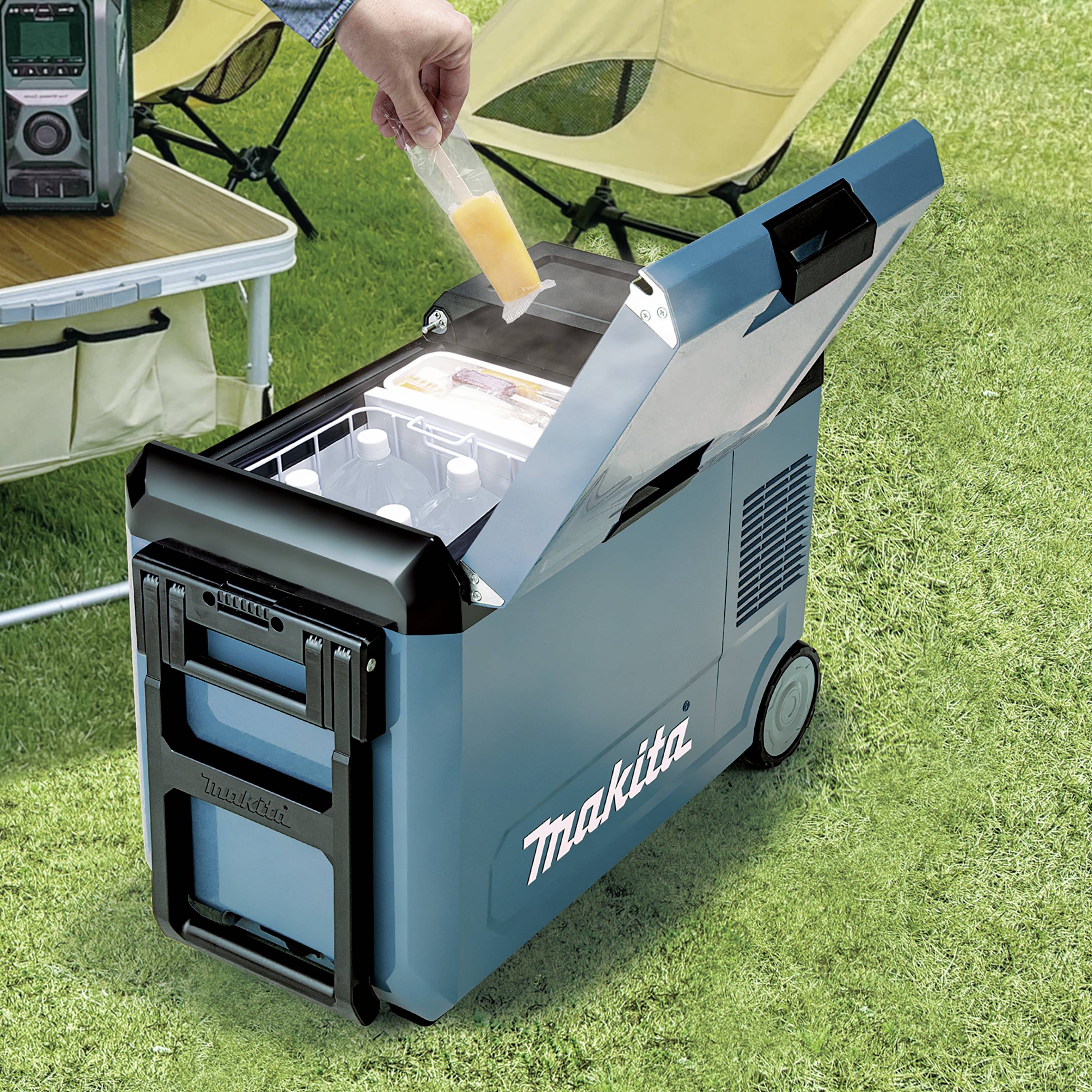 Een persoon vult een ijsblokjeshouder in een geopende blauwe Makita-koelbox, die op gras naast een campingtafel staat.
