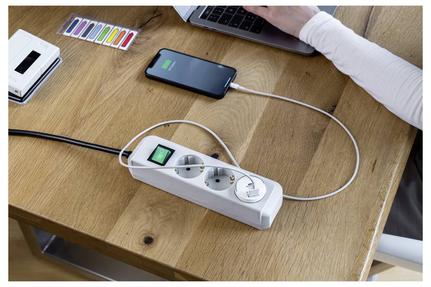 Smartphone oplaadbaar via een stekkerdoos naast een laptop op een houten bureau, met een persoon die in de buurt werkt en kantoorartikelen op de achtergrond.