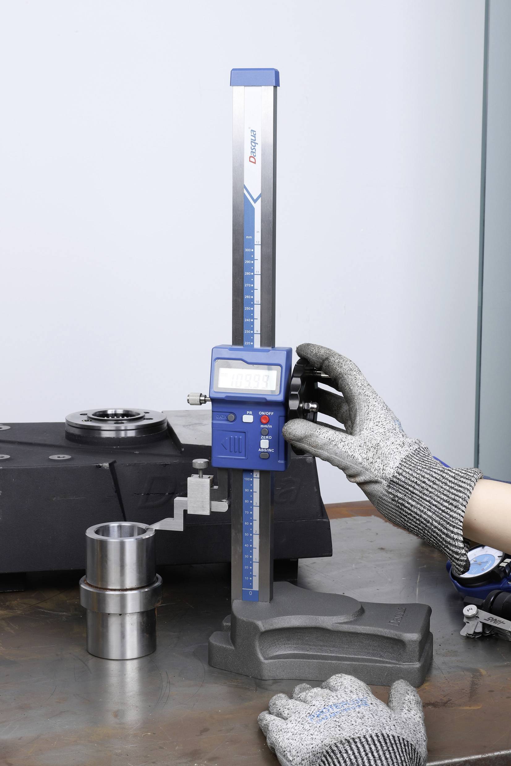 Een persoon meet een metalen object op een tafel met een digitale hoogtemeter. Draagt werkhandschoenen.