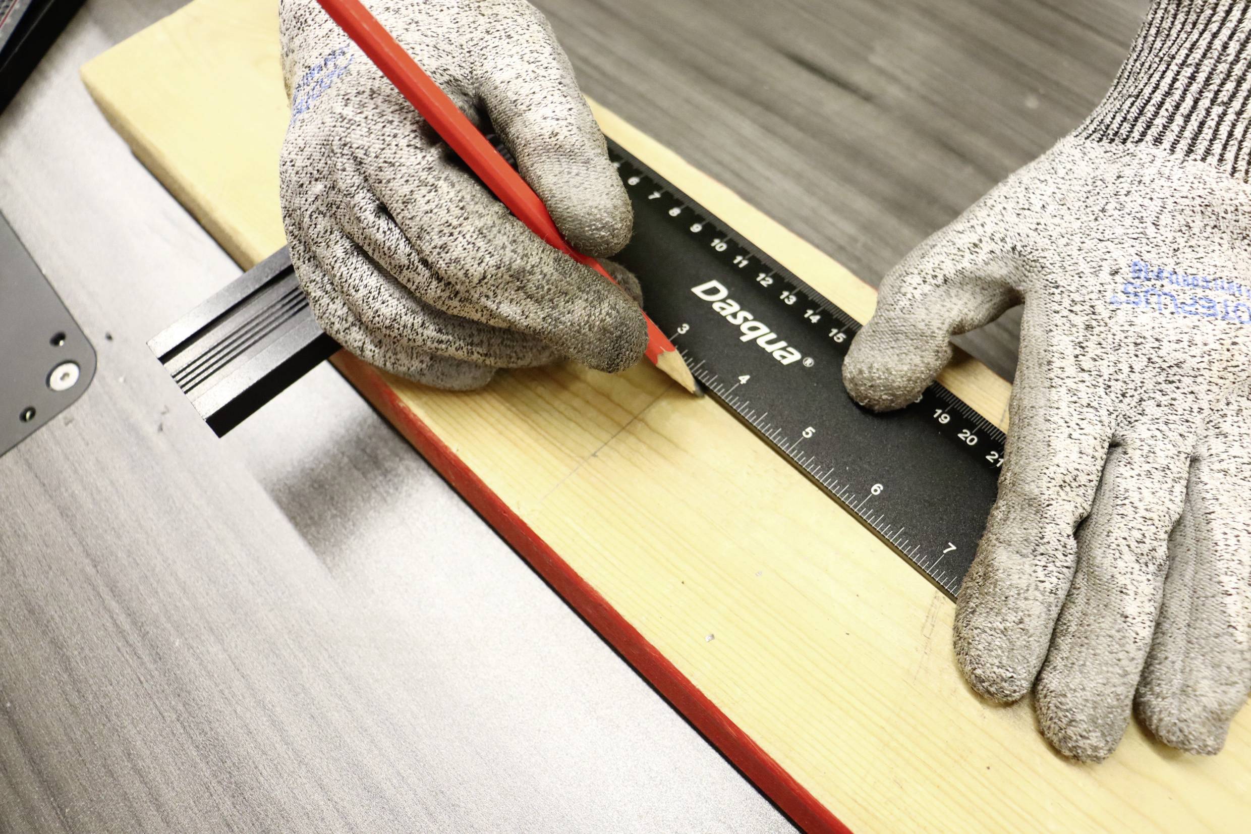 Een persoon met grijze handschoenen markeert een houten plank met een rode potlood en een zwarte liniaal op een tafel.