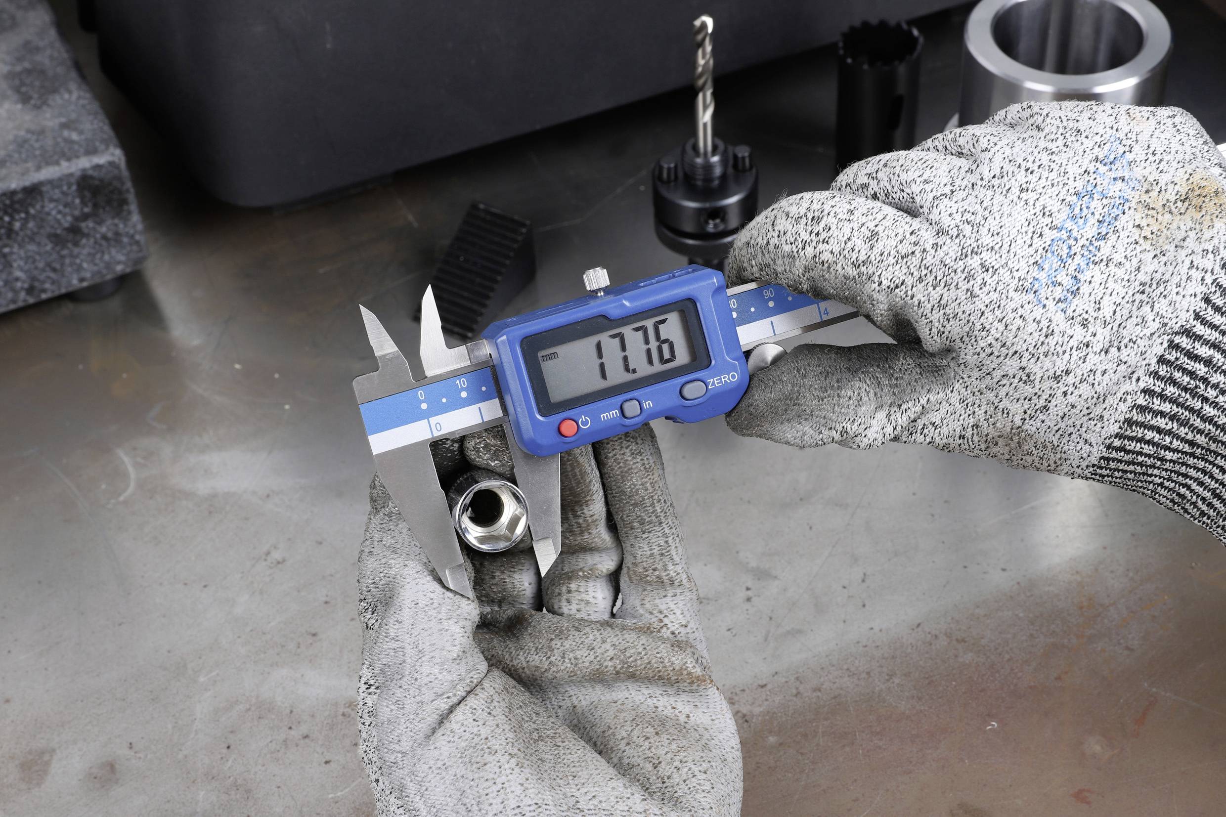 Een persoon draagt handschoenen en meet de diameter van een metalen stuk met een digitale schuifmaat. Het scherm toont '17,76'.