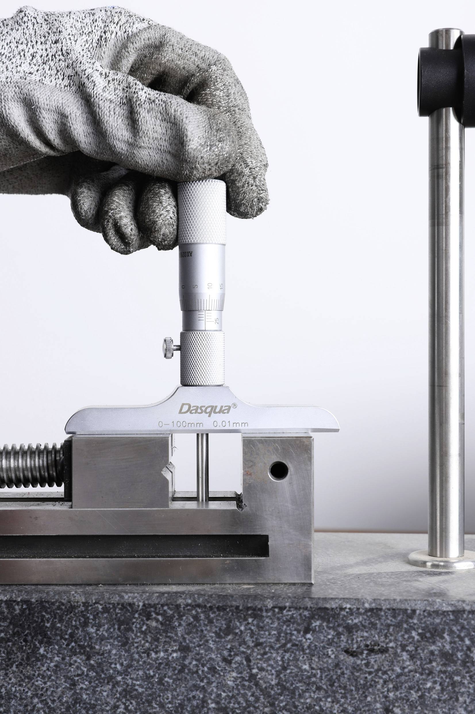 Een hand in een handschoen houdt een hoogtemeter vast die een metalen onderdeel meet, gemonteerd op een werkbank.