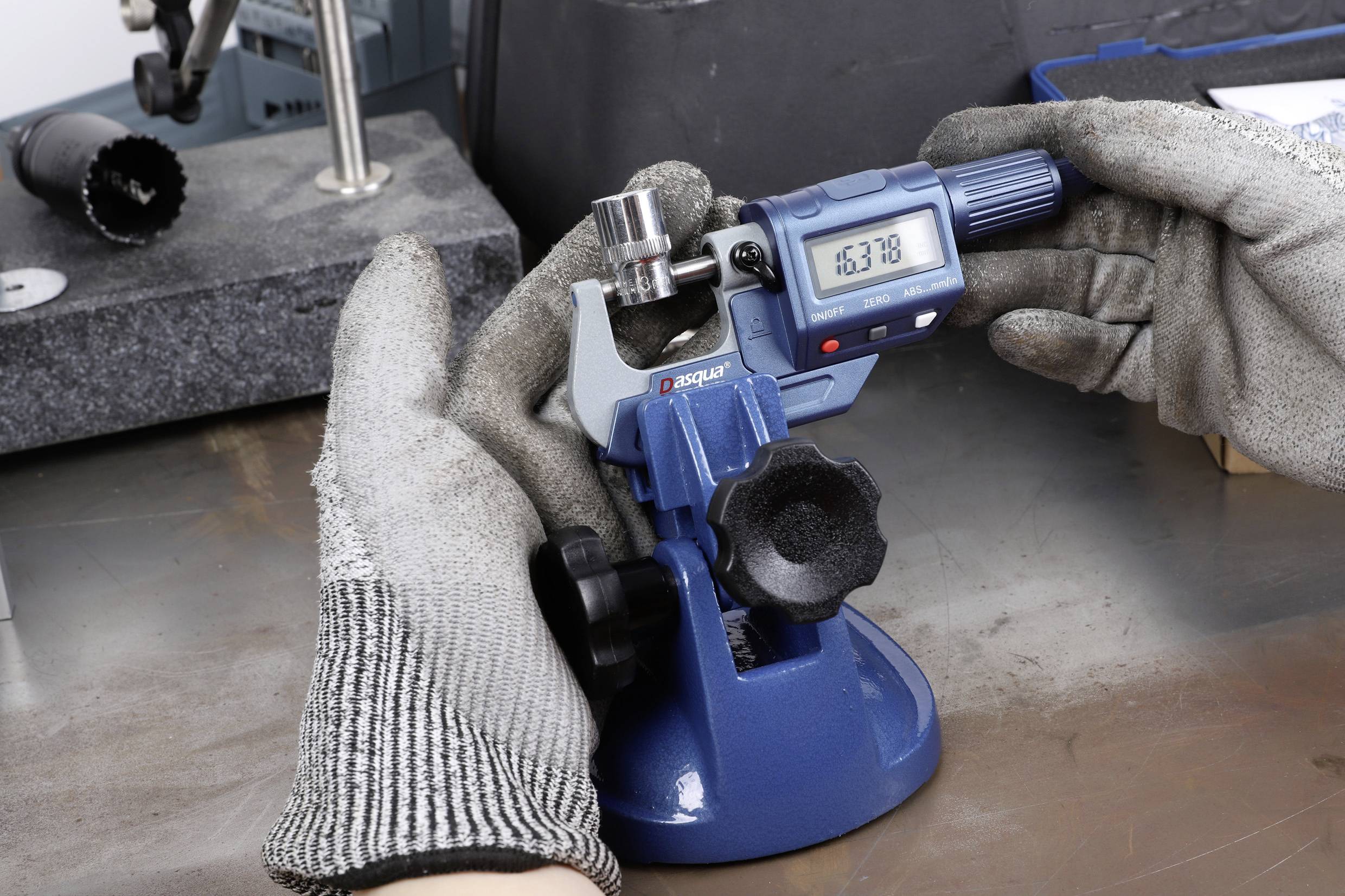 'Close-up van handen in grijze handschoenen die een blauw micrometer aanpassen op een rechthoekige metalen voorziening op een tafel.'