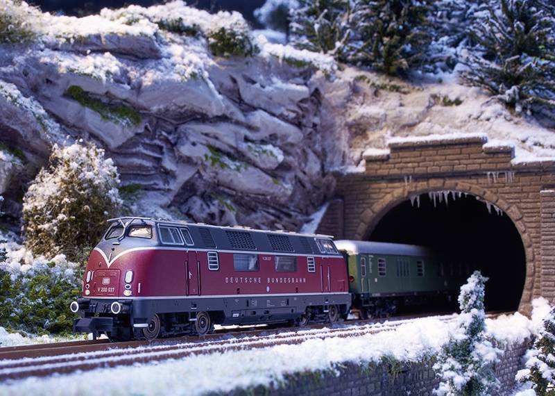 Een modeltreintje rijdt uit een tunnel in een besneeuwde landschap. De locomotief is rood, met 'Deutsche Bundesbahn' erop geschreven.