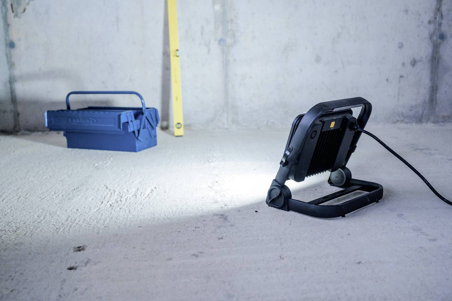 Een draagbare werklamp verlicht een bouwplaats met een betonnen vloer, ernaast staat een blauwe gereedschapskist en een gele waterpas.