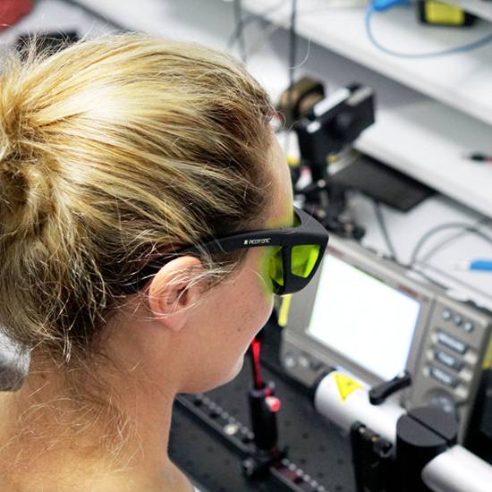 Een persoon met een veiligheidsbril werkt aan een lasertoestel in een laboratorium, omringd door technische apparatuur en monitors.