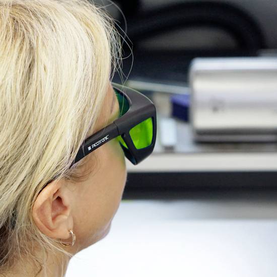 Een persoon met blond haar draagt een veiligheidsbril met groene glazen. De persoon bevindt zich kennelijk in een laboratoriumomgeving.
