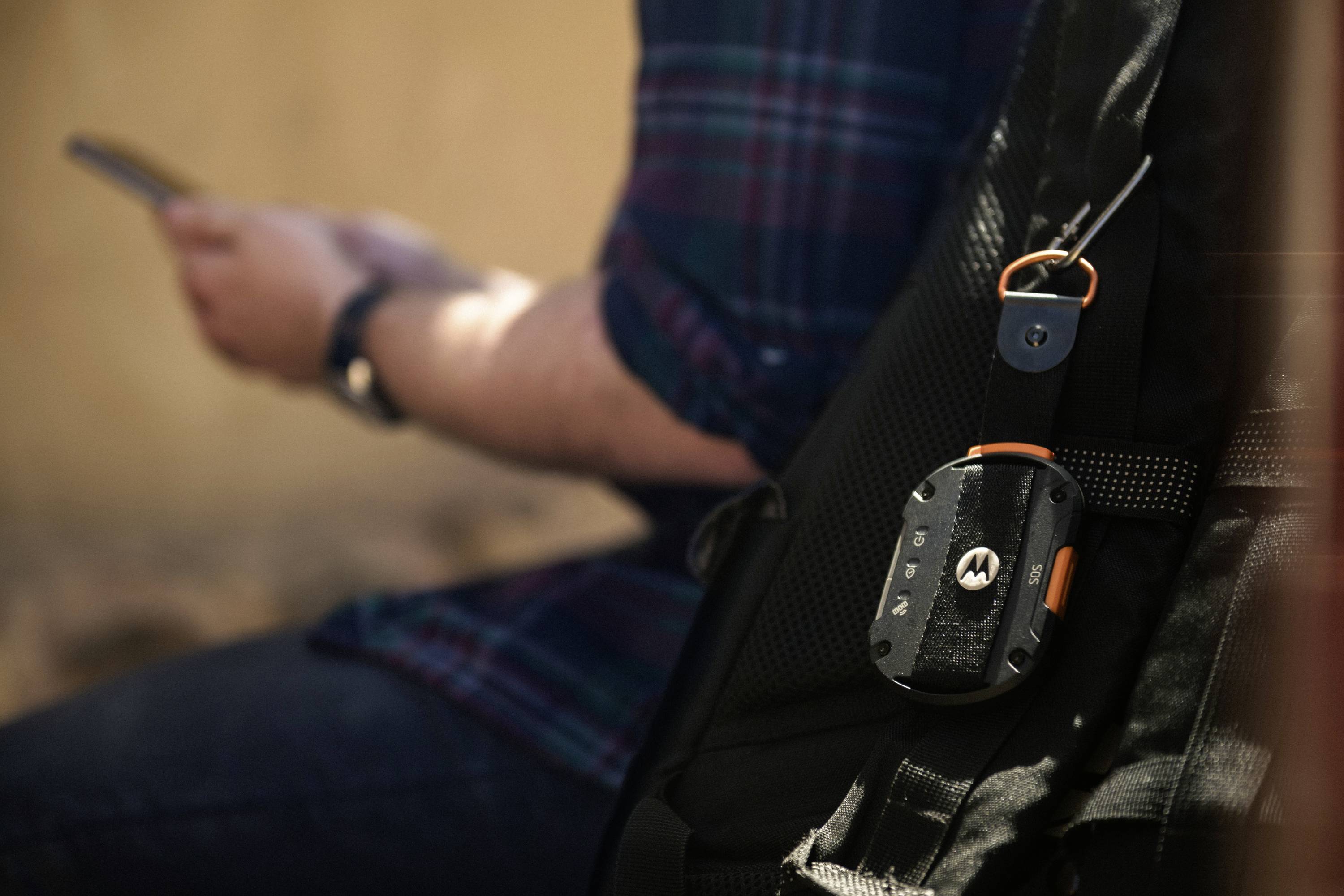 Een persoon zit met een smartphone in de hand. Aan de rugzak hangt een apparaat met het Motorola-logo. De sfeer oogt ontspannen.