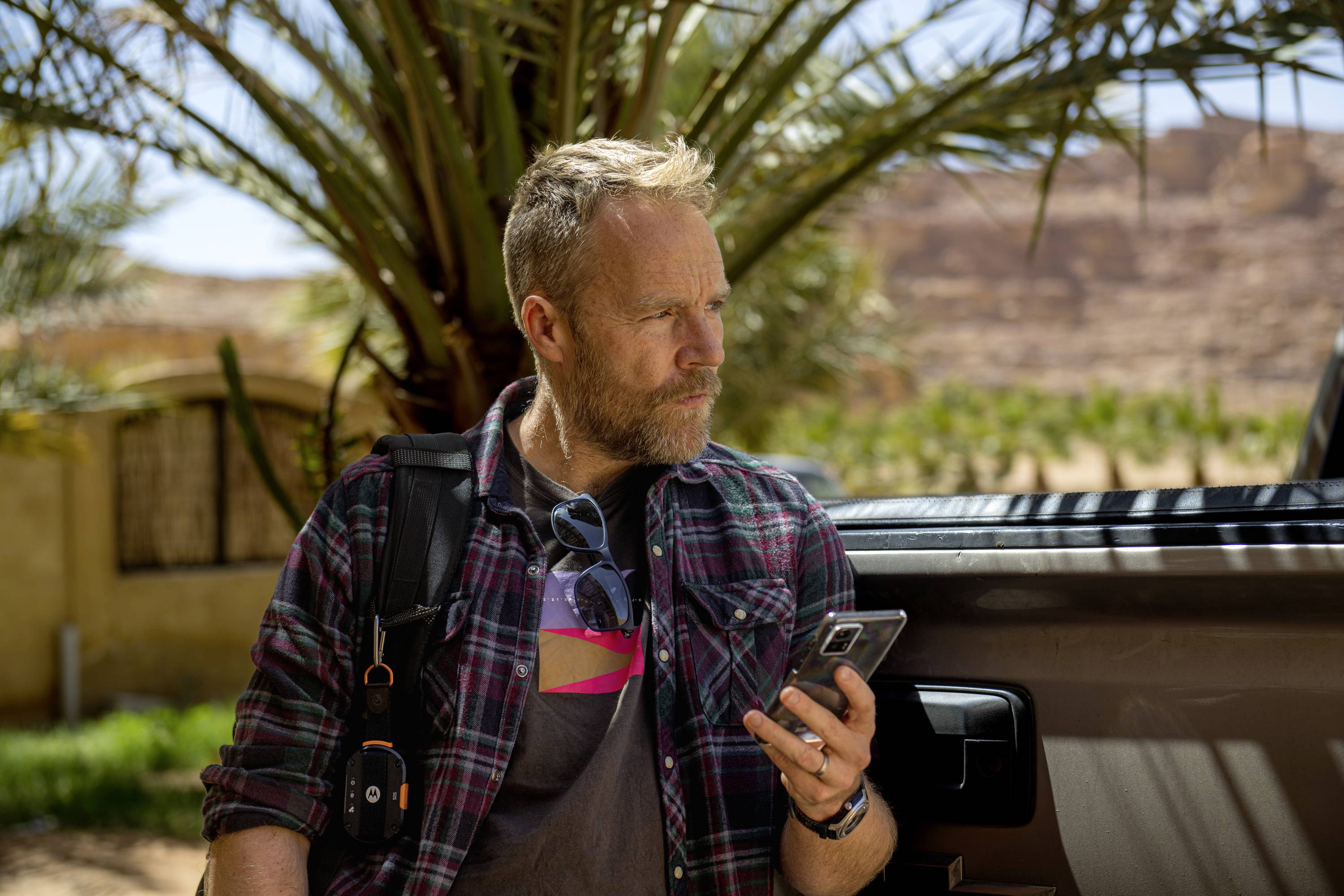 Een persoon met een baard houdt een smartphone vast en staat naast een auto. Op de achtergrond staat een palmboom en een woestijnlandschap.