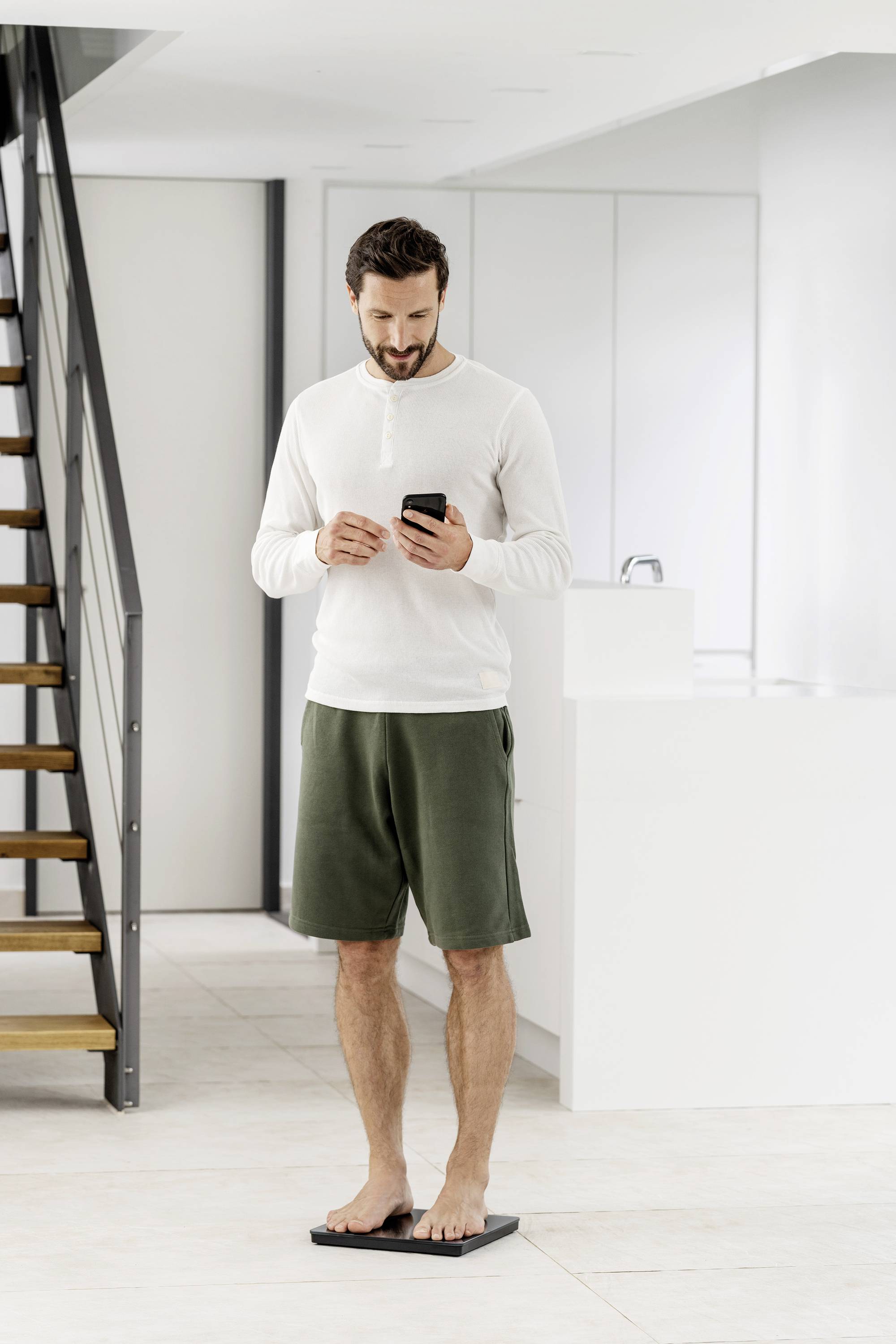 Een man staat op een weegschaal in een moderne, lichte ruimte en kijkt naar zijn smartphone.