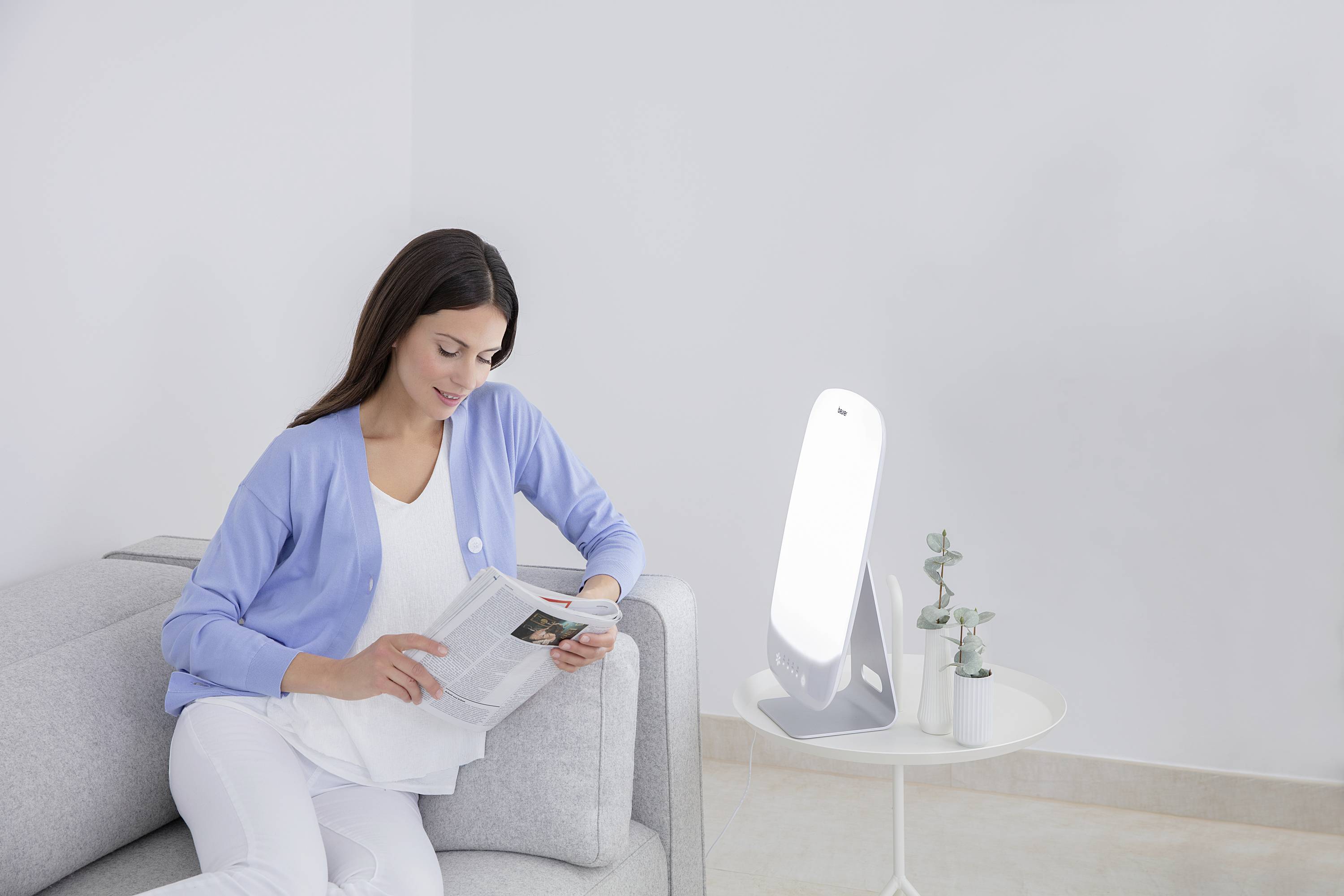 Een vrouw zit op een bank en leest een boek. Naast haar staat een lamp op een klein tafeltje. De omgeving is licht en eenvoudig.