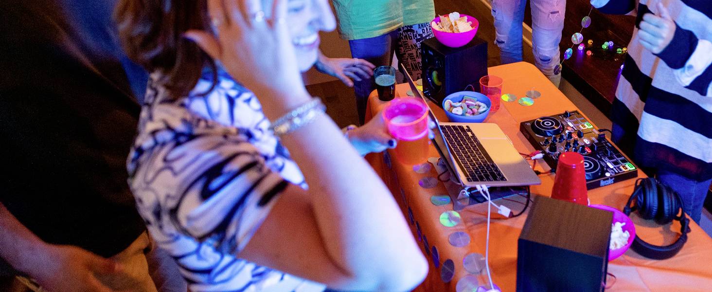 Iemand danst naast een tafel met een laptop en een DJ-booth, omringd door drankjes en hapjes. De sfeer is vrolijk en uitbundig.