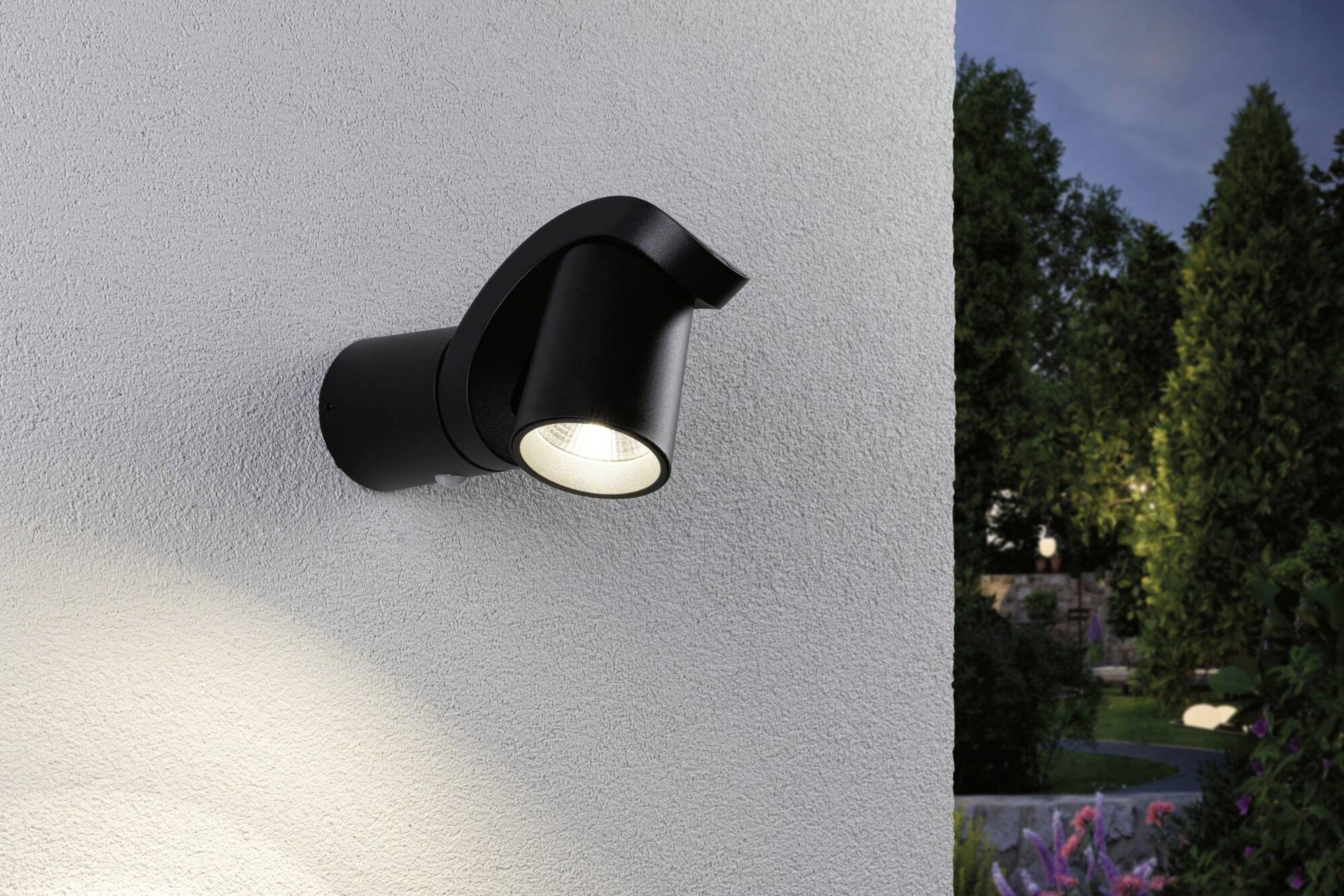 Buitenwandlamp in modern design verlicht een muur bij schemering, omgeven door planten en tuin.