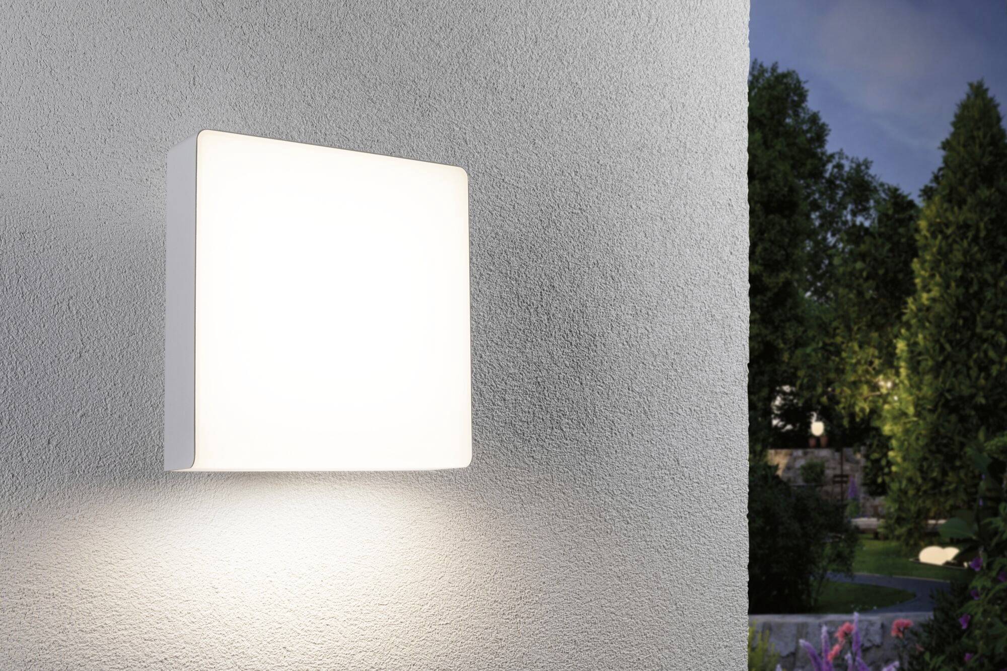 Een lichtgevende, vierkante buitenlamp aan een gevel die bij het invallen van de schemering een tuin met bomen en struiken verlicht.