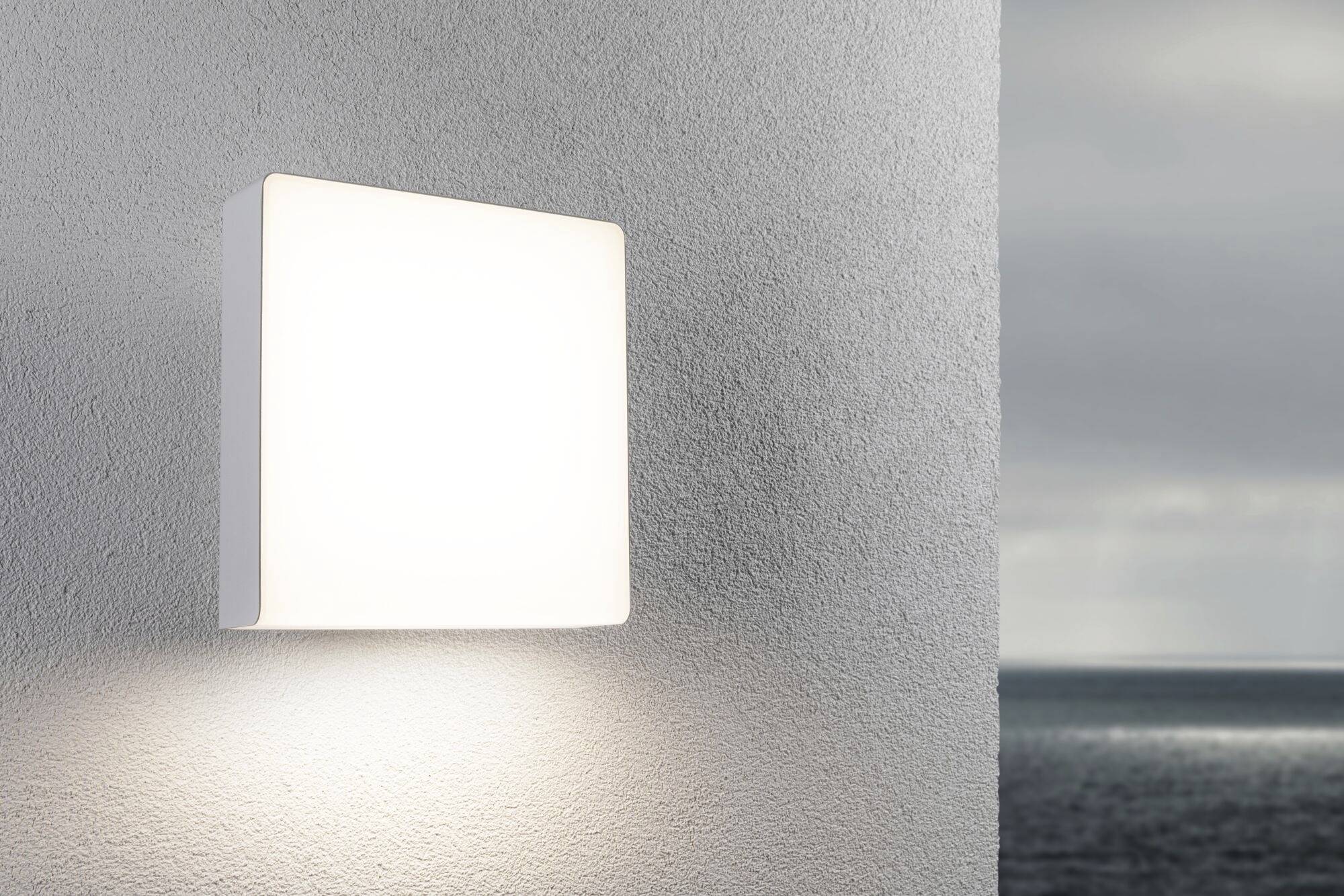 Een vierkante, moderne lamp schijnt tegen een grijze muur met uitzicht op een grijze, bewolkte zee.