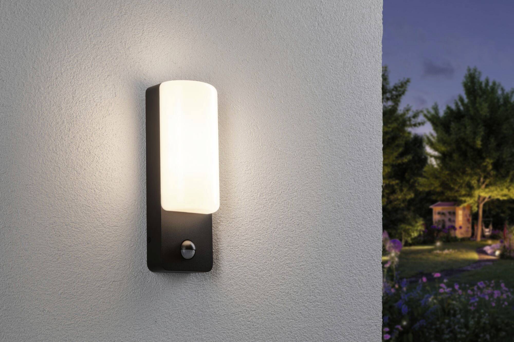 Een moderne buitenmuurlamp met een rechthoekige witte kap verlicht een muur bij het invallen van de avond. Op de achtergrond is een tuin zichtbaar.