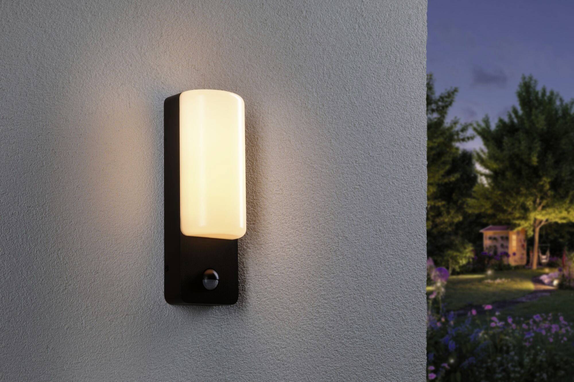 Wandlamp schijnt aan een muur, naast een tuin bij schemering met bomen en een klein huisje op de achtergrond.