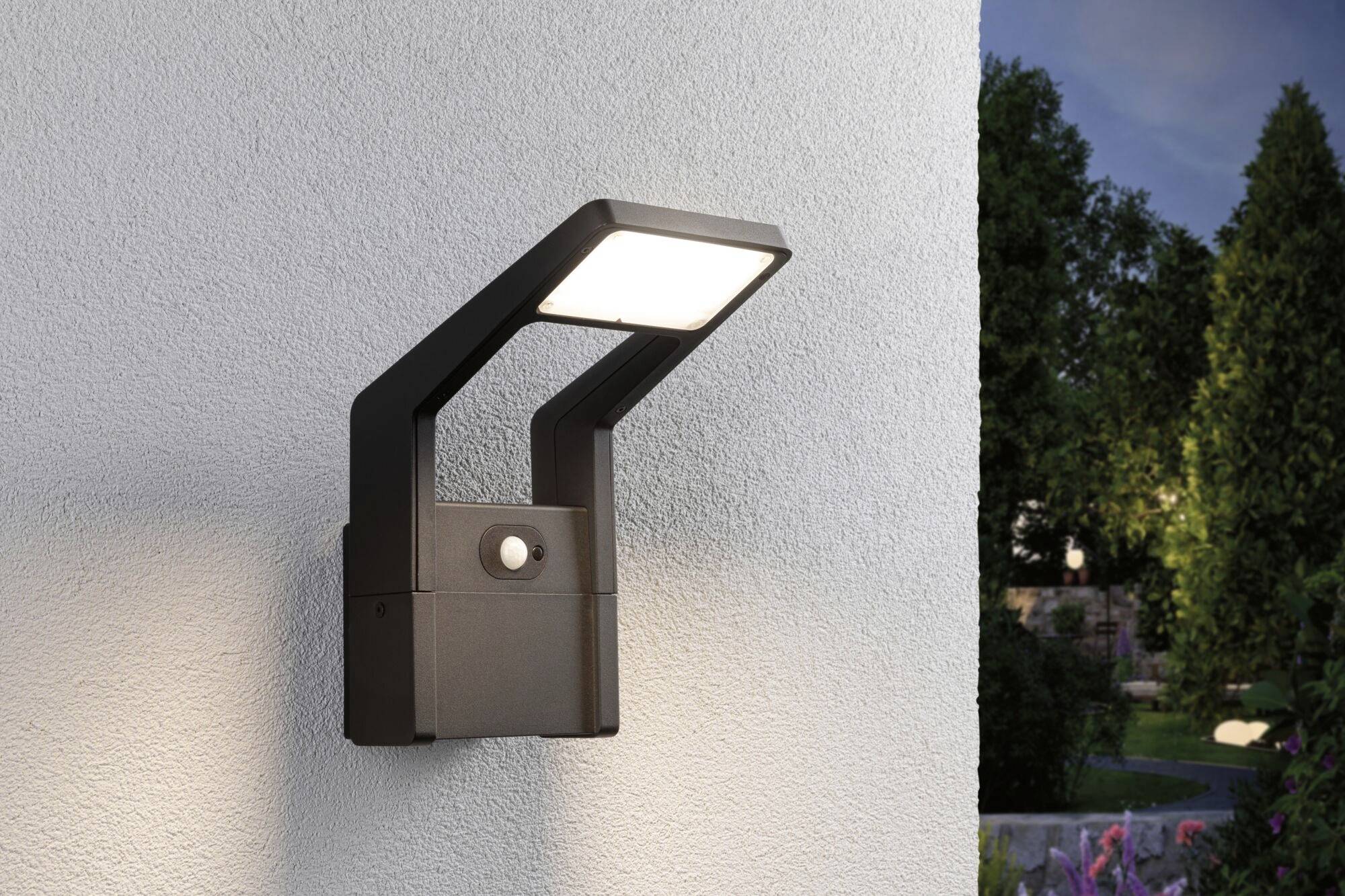 Buitenlamp met bewegingssensor aan een buitenmuur, verlicht het gebied bij schemering; tuin zichtbaar op de achtergrond.