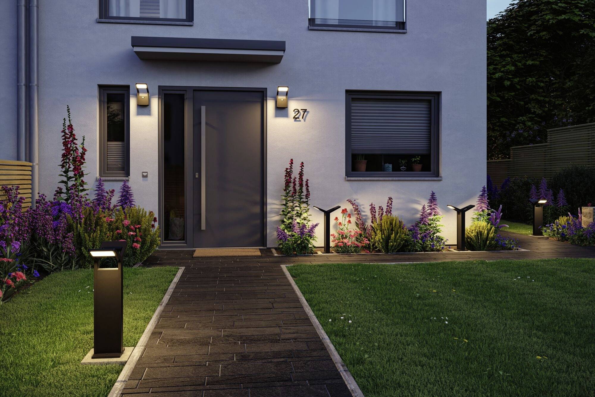 Een modern huis met een grijze deur en huisnummer '27' bij het invallen van de avond, omgeven door kleurrijke bloemen en verlichte tuinpaden.