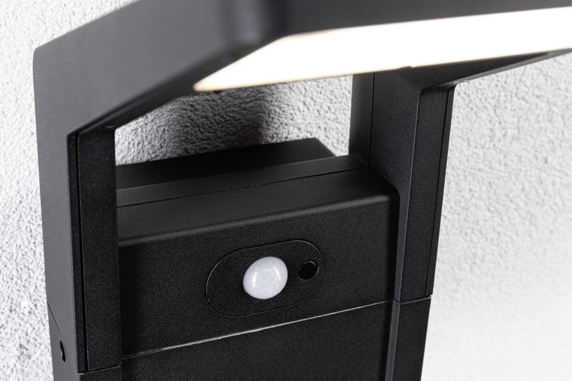 Een zwarte buitenlamp met ingebouwde bewegingssensor aan een grijze muur, die van boven naar beneden schijnt.