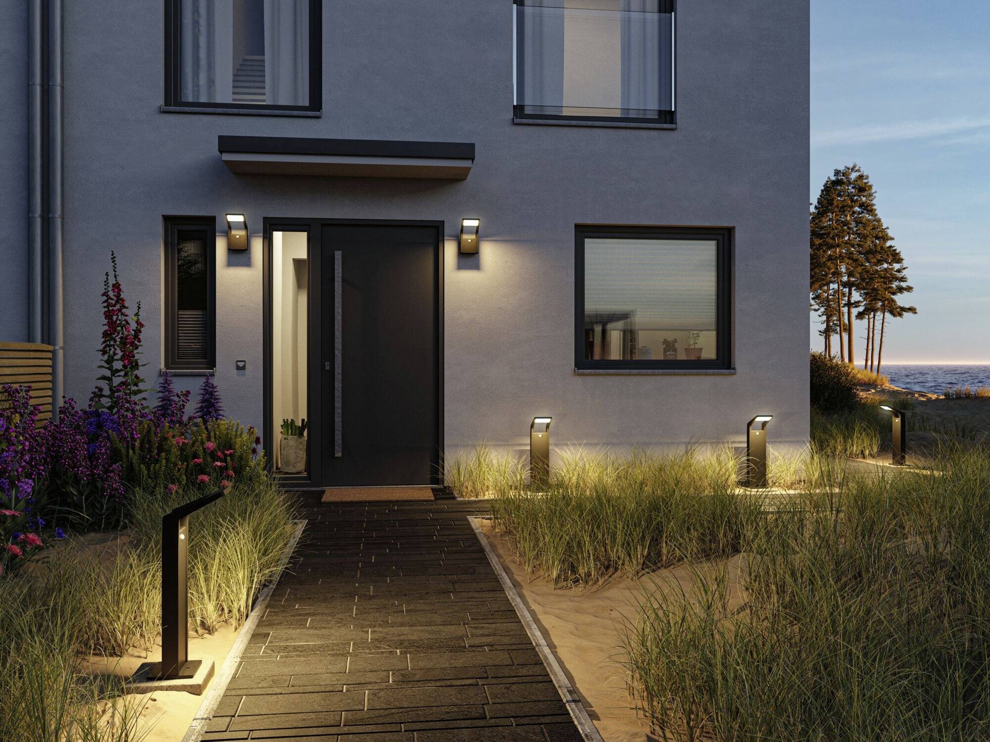 Toegang tot een modern huis bij schemering. Een verlicht pad met gras en buitenverlichting beheerst het beeld, met zee op de achtergrond.