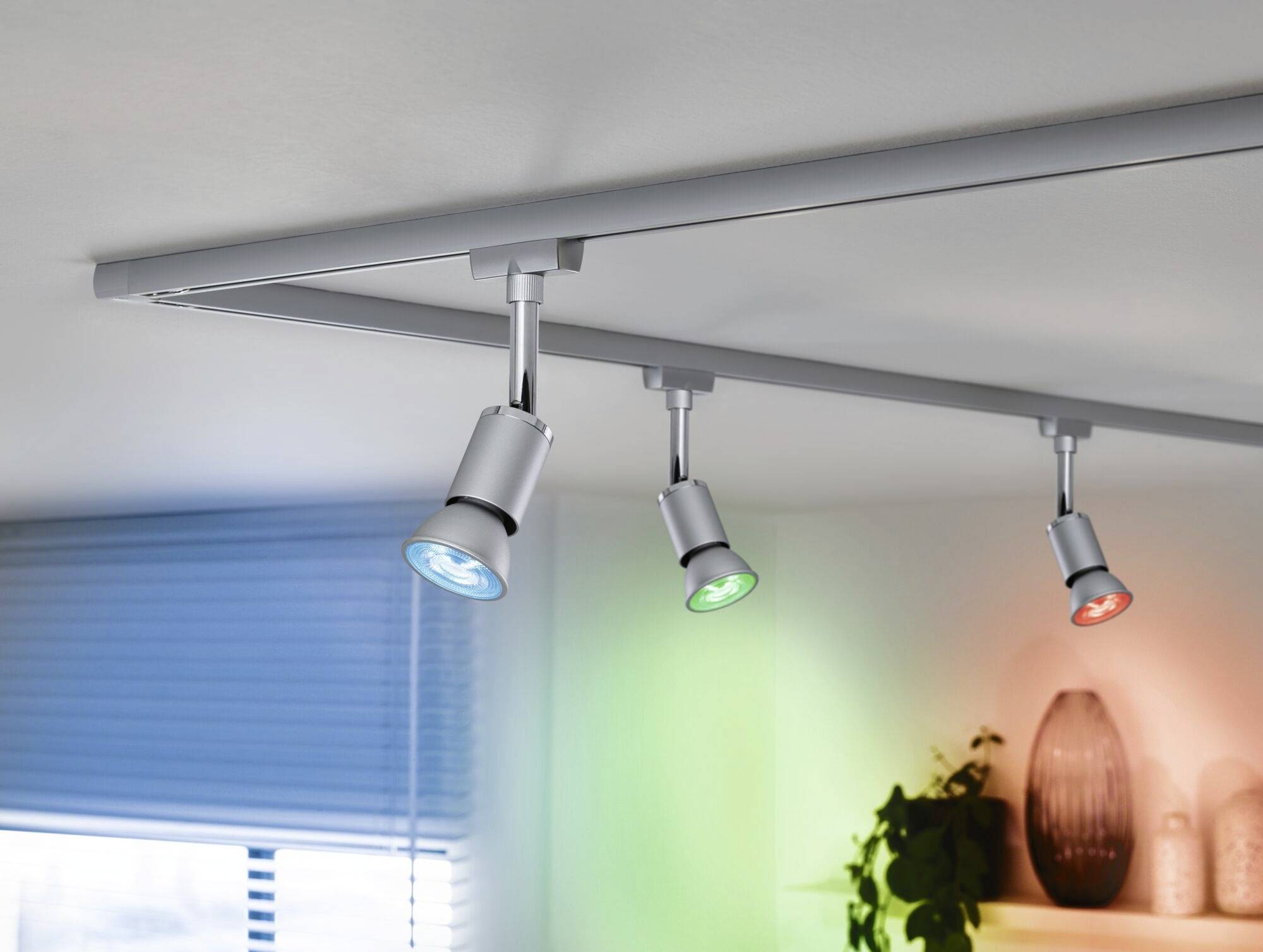 Plafondlampen met gekleurd licht (blauw, groen, rood) schijnen op een witte plafond. Planten en vazen zijn zichtbaar op de achtergrond.