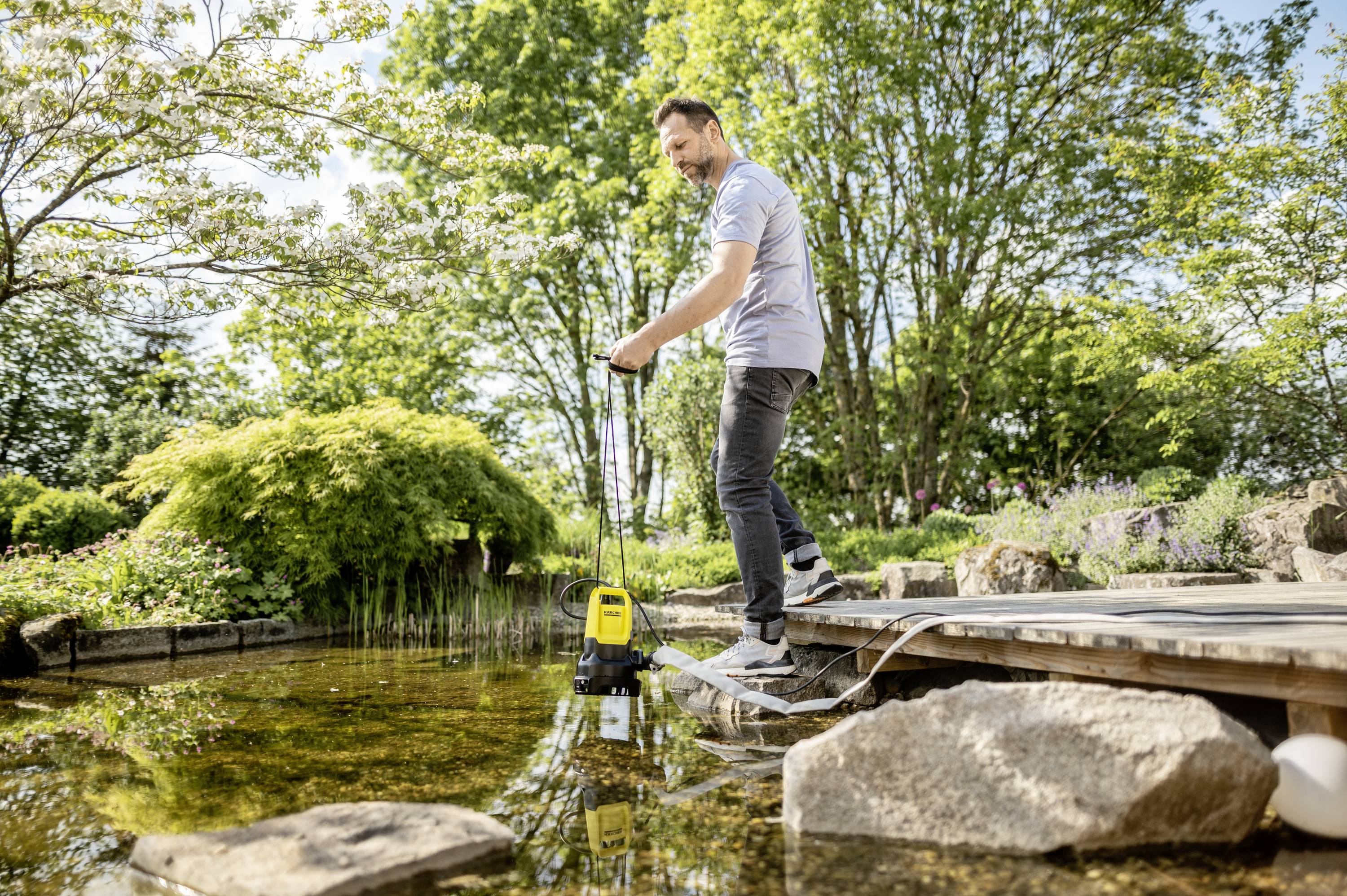 Een man staat op een houten steiger en houdt een waterpomp boven een vijver. Bomen en planten zijn te zien op de achtergrond.