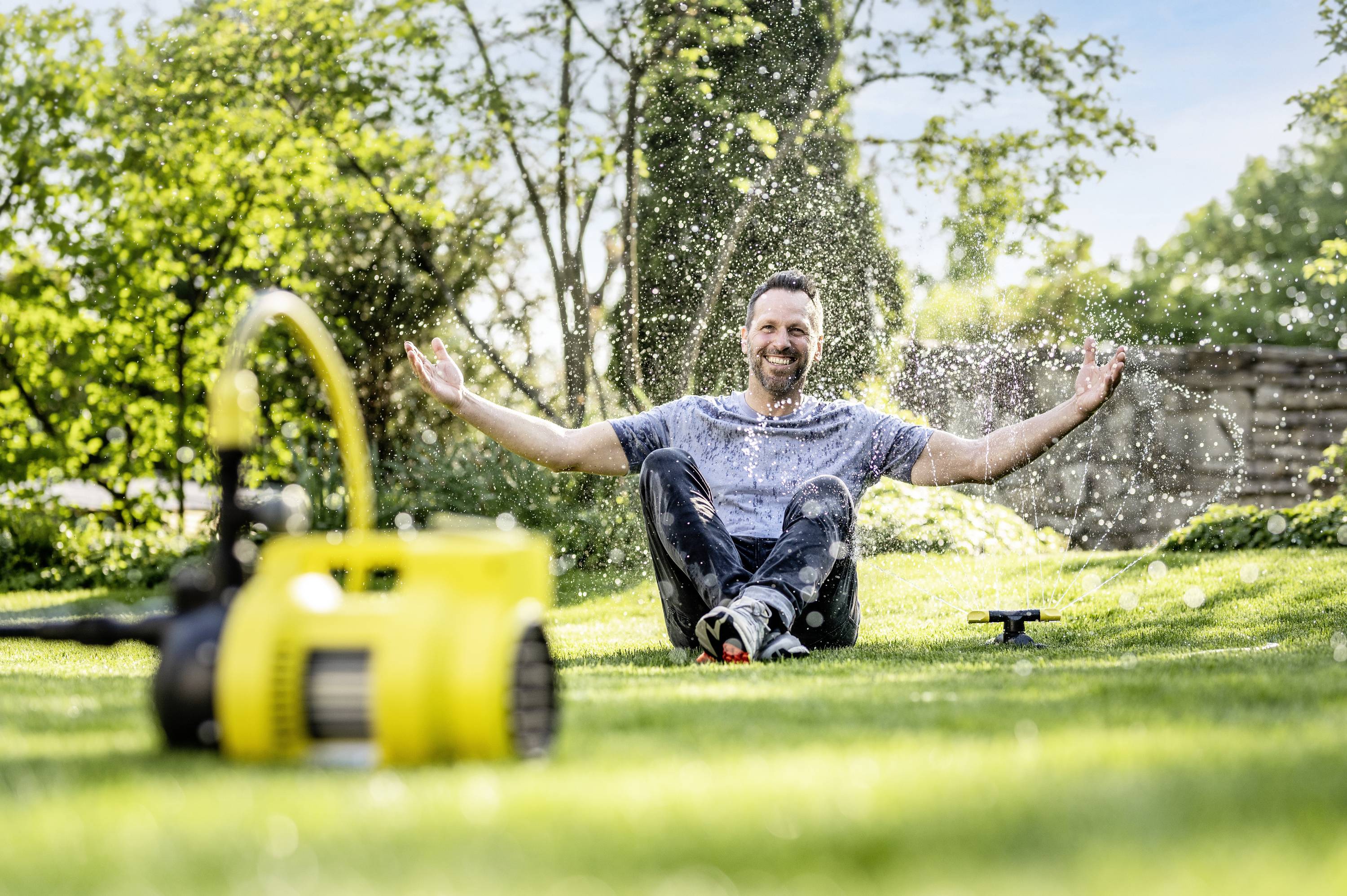 Een man zit lachend op pas besproeide grasmat, omringd door rondspattend water, met op de achtergrond een zomerse tuinsfeer.