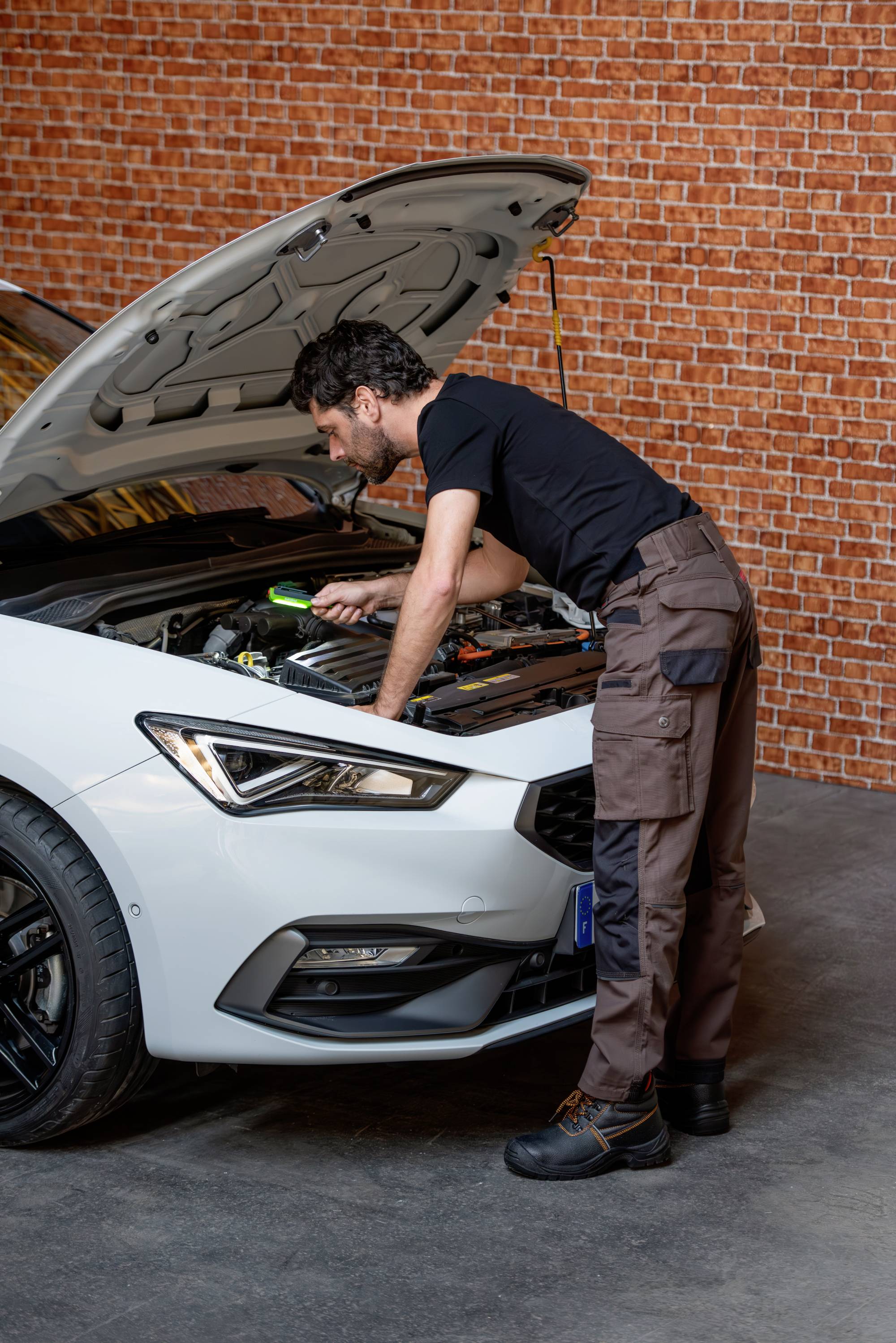 Een monteur repareert de motor van een witte auto in een werkplaats. De motorkap staat open, en de monteur werkt geconcentreerd.