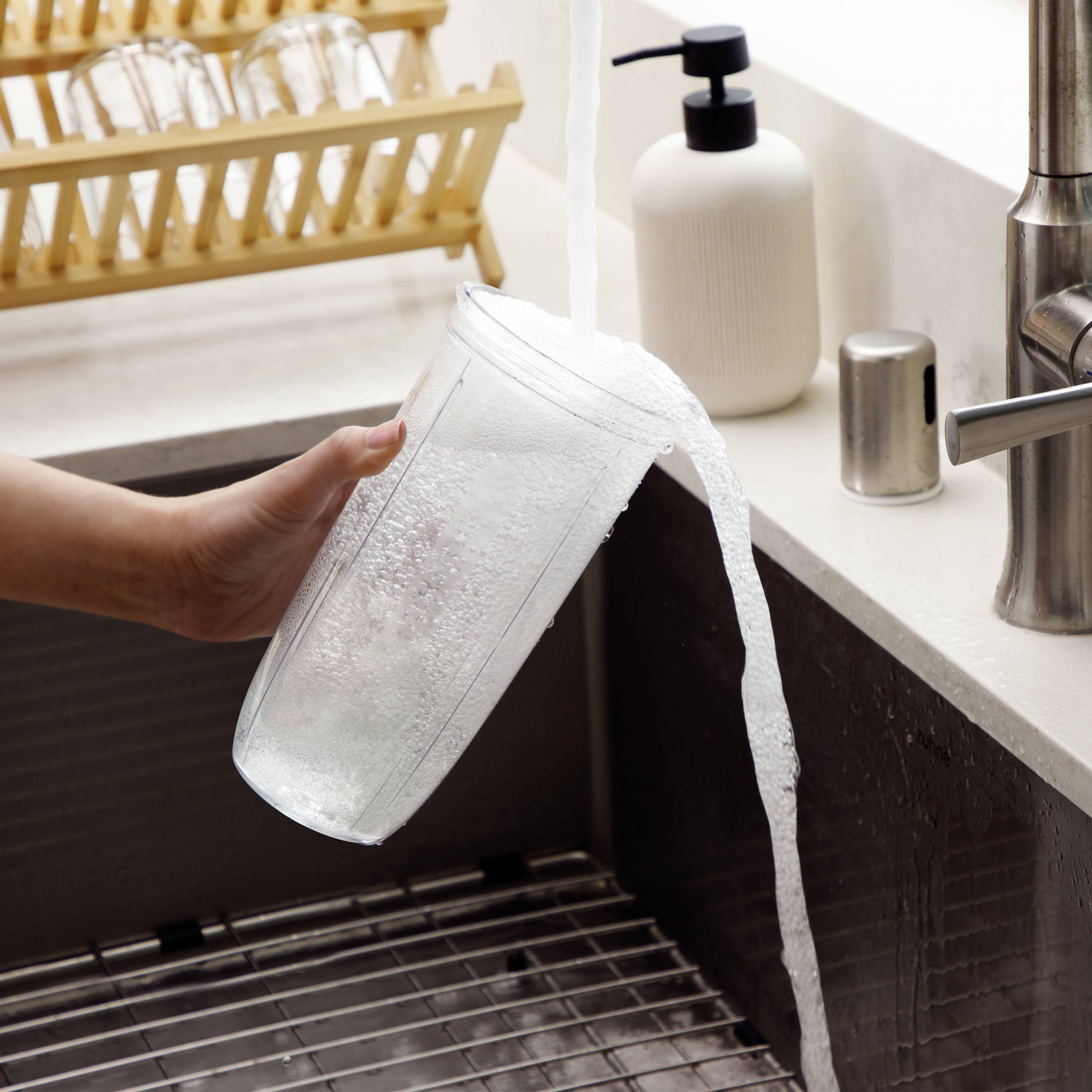 Een hand wast een drinkglas onder stromend water in een gootsteen. Op de achtergrond staan een zeepdispenser en een afwaasrek.