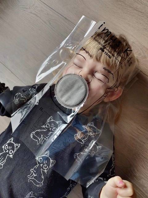 Een kind ligt op een houten vloer en draagt een transparant masker met een rond filter. Het draagt een trui met een kattenpatroon.