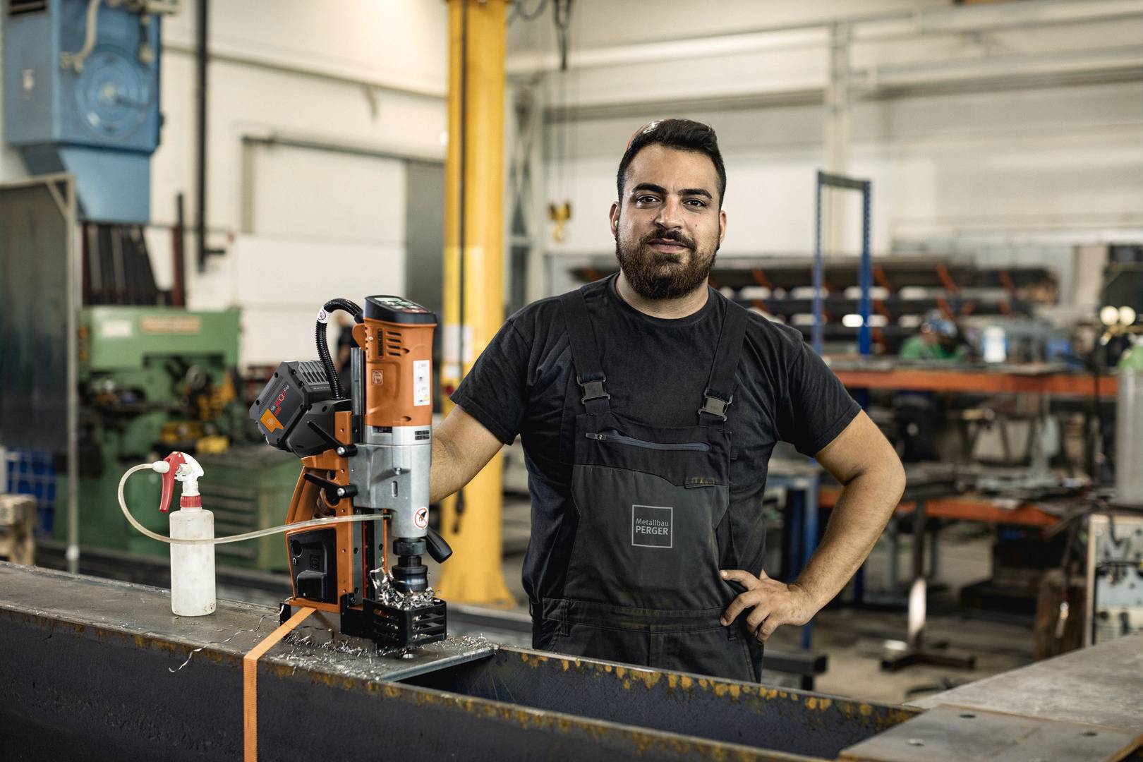 Een man staat in een werkplaats naast een boormachine. Hij draagt werkkleding en lacht naar de camera. Op de achtergrond zijn gereedschappen en machines te zien.