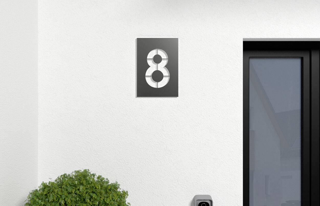Gevel met het huisnummer 8 op een bord. Rechts ernaast een zwarte deur. Links onderaan is een groene struik zichtbaar.