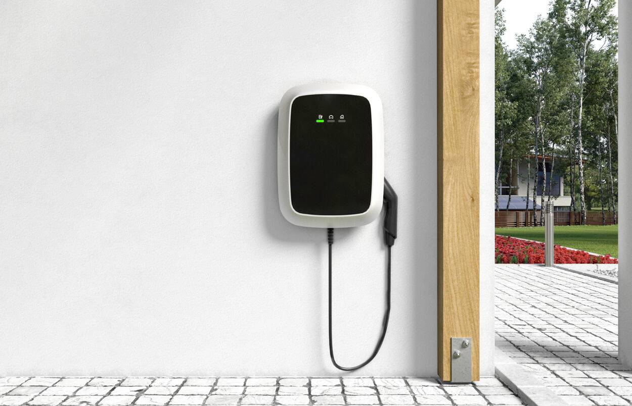 Laadpunt voor elektrische auto's gemonteerd aan een witte muur, met groen licht. Kabel hangt naar beneden. Tuin zichtbaar op de achtergrond.