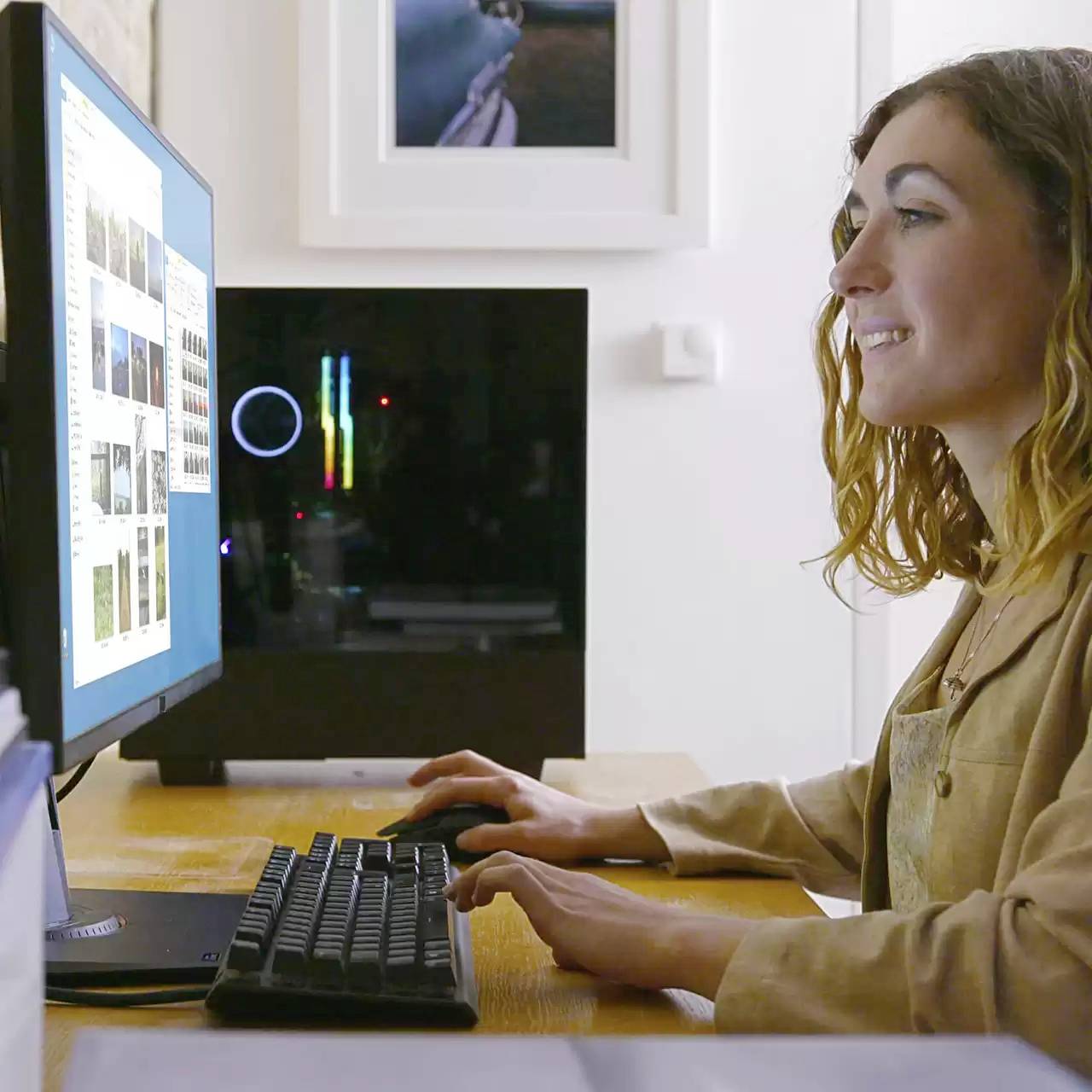 Een vrouw zit lachend aan een bureau en bekijkt inhoud op een computerscherm. Op de achtergrond is een fotolijst zichtbaar.