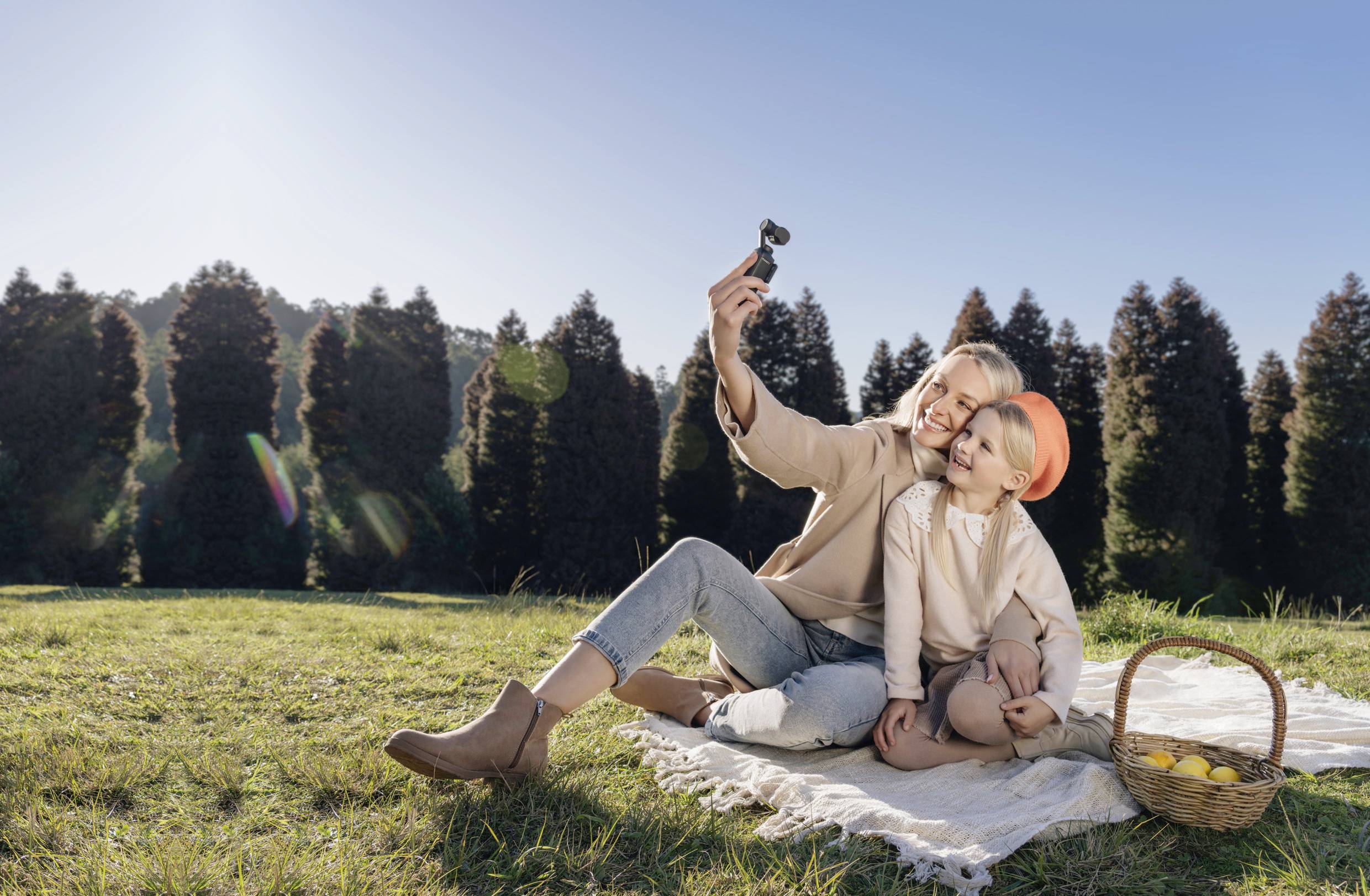 Een vrouw en een kind zitten op een deken buiten en maken een selfie. Een picknickmand met appels staat ernaast. Op de achtergrond zijn bomen.