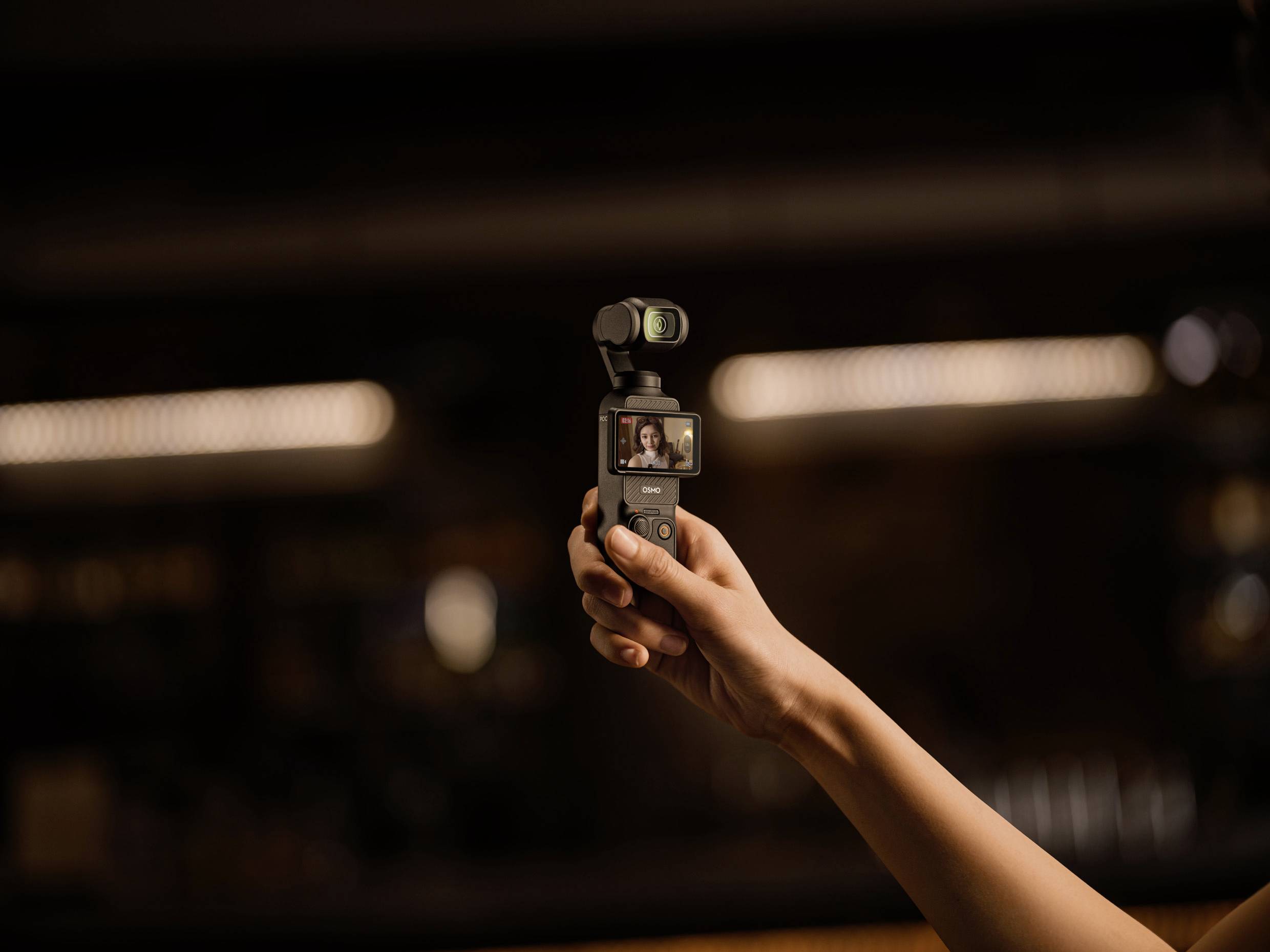Een hand houdt een kleine camera met een scherm dat het beeld van een persoon toont. De omgeving is donker met onscherpe lichten op de achtergrond.