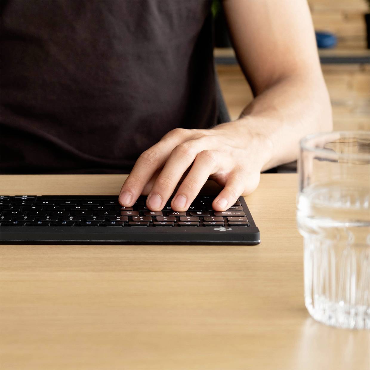 Een hand tikt op een zwart toetsenbord. Naast het toetsenbord staat een glas water op een houten tafel.