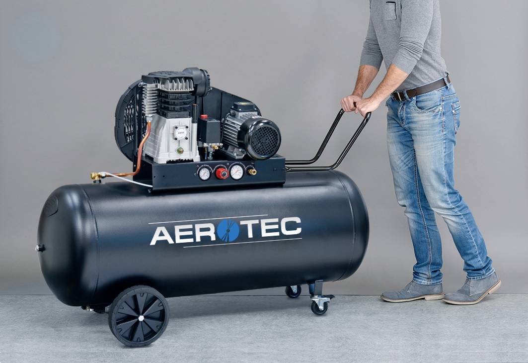 Een persoon duwt een grote, zwarte luchtkompressor op wielen. Op de kompressor staat 'AEROTEC'.