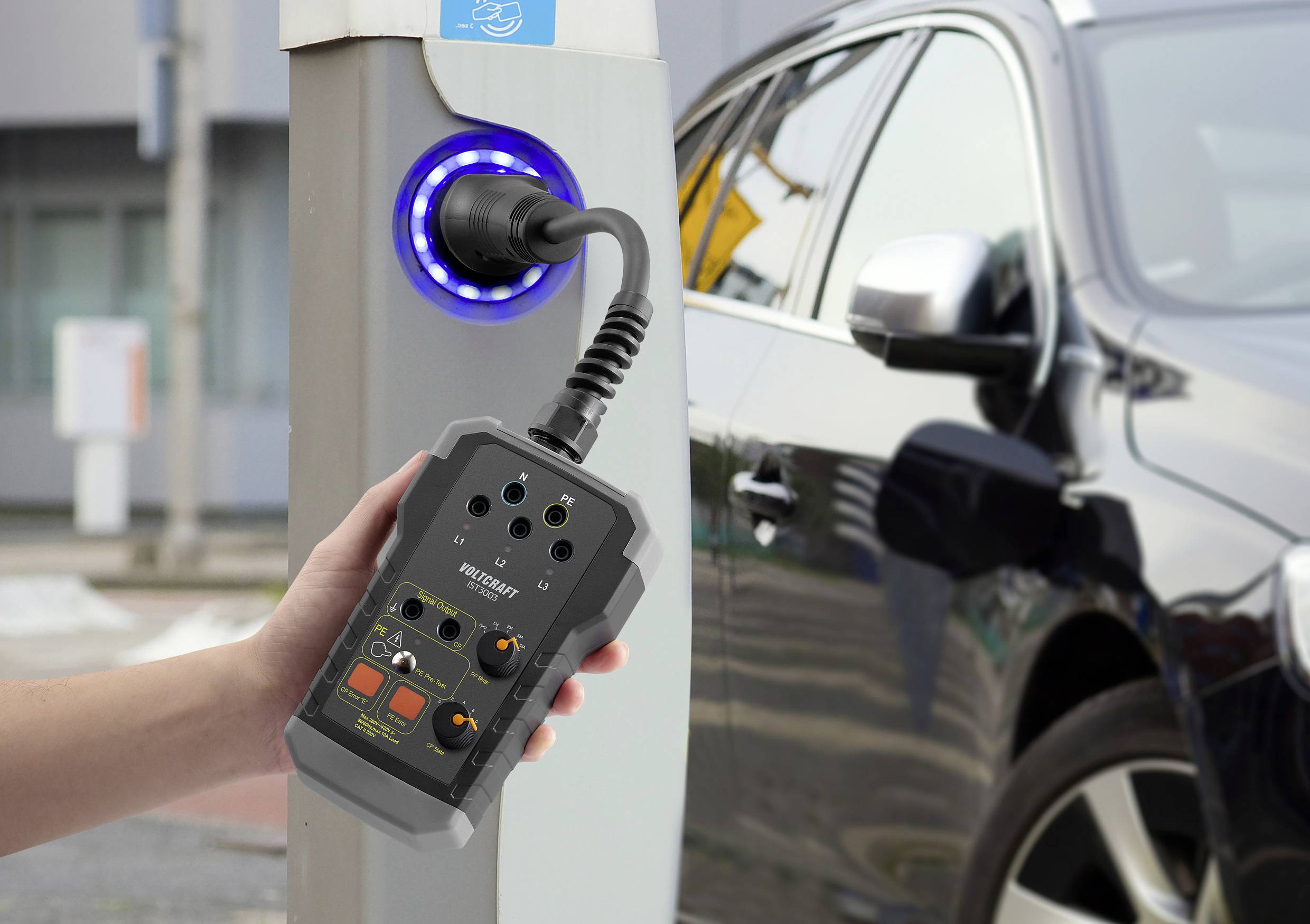 Een hand houdt een meetapparaat vast dat is aangesloten op een laadstation terwijl een auto wordt opgeladen.