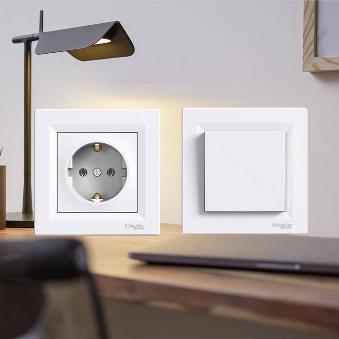Twee witte schakelaars aan een houten wand, verlicht door een moderne bureaulamp. Op de achtergrond zijn delen van een bureau zichtbaar.