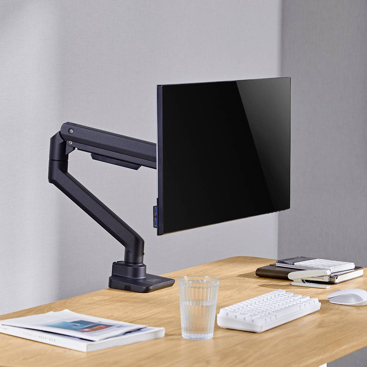 Een moderne bureau met een computerbeeldscherm op een scharende arm, een toetsenbord, een muis, een glas water en notitieboeken.