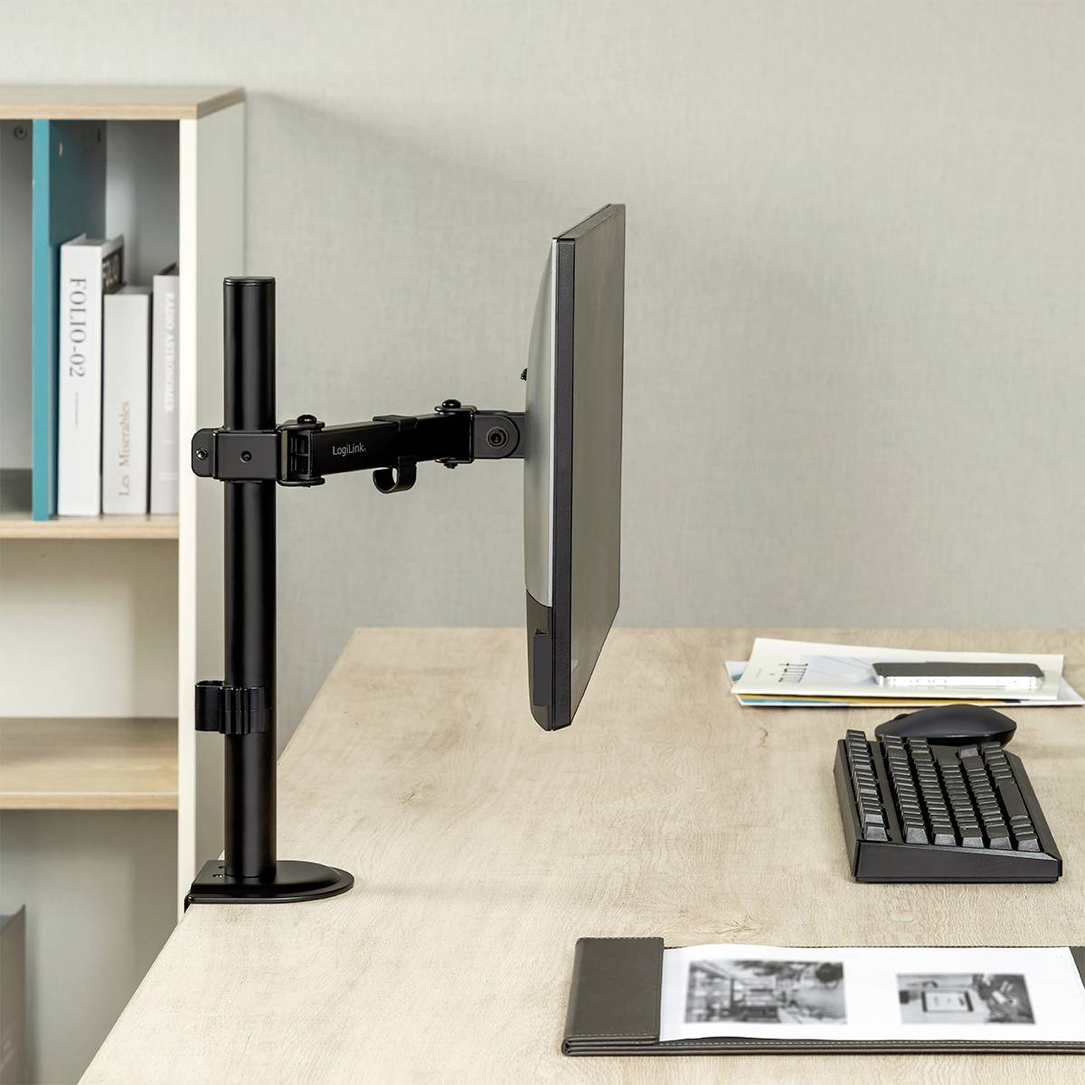 Een kantoorwerkplek met een monitor aan een verstelbare arm, computermuis, toetsenbord en notitieboek op een licht houten bureau. Op de achtergrond een plank met boeken.