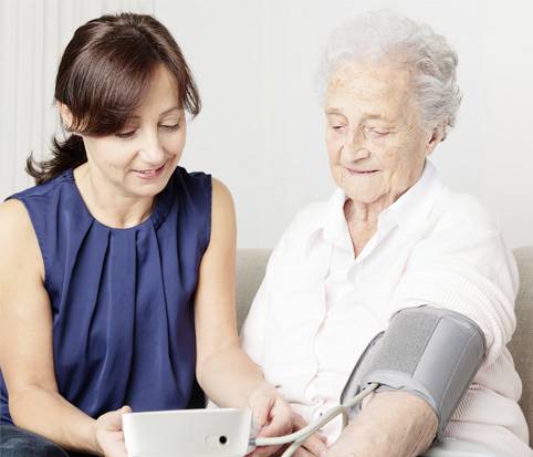 Een jonge vrouw meet de bloeddruk van een oudere vrouw. De oudere vrouw draagt een bloeddrukmeter aan haar arm, beiden glimlachen.
