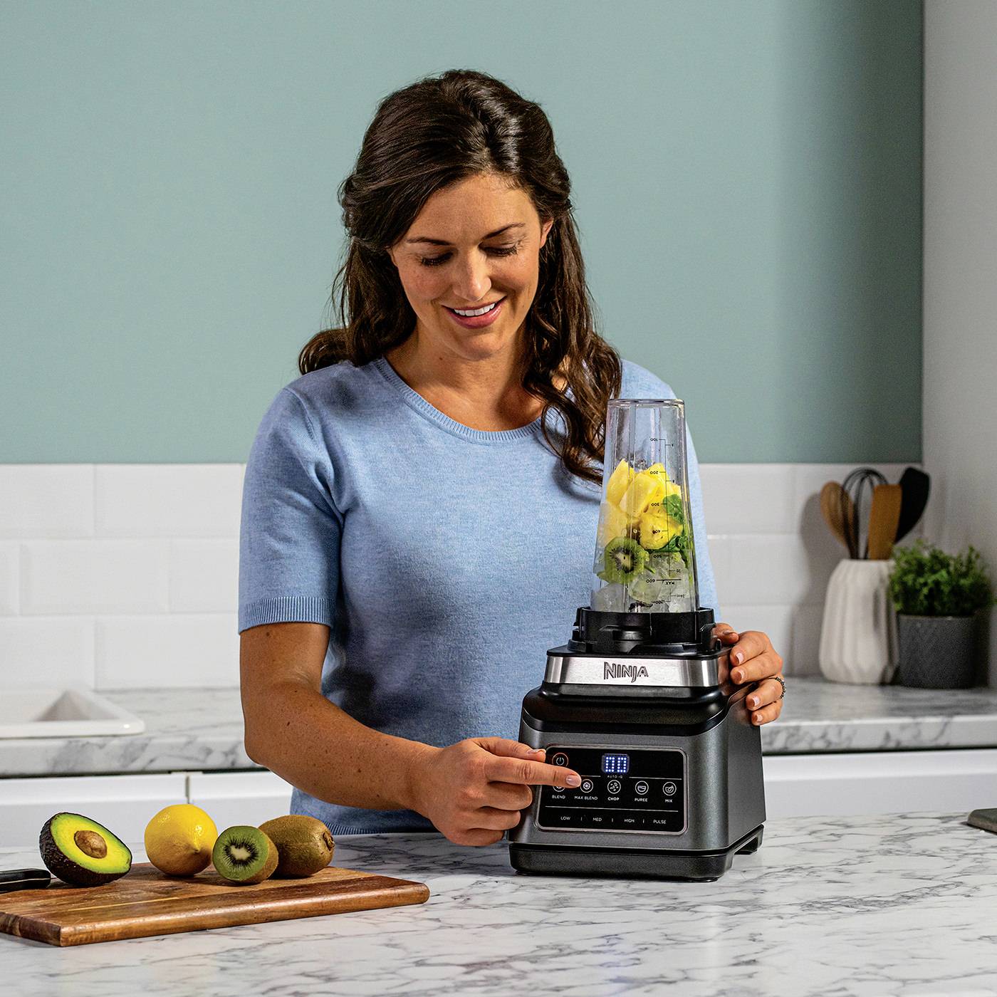Een vrouw gebruikt een blender op een aanrecht om fruit, zoals kiwi en ananas, te mengen. Op het aanrecht liggen gesneden stukken fruit.